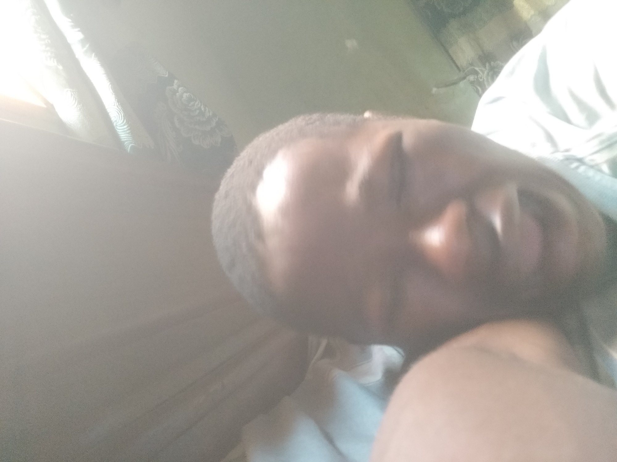 A person with dark skin and short dark hair is lying down, with the right side of their face visible and mouth slightly open. The person appears to be resting indoors. A white and floral curtain is visible in the upper left corner. The environment appears to be a bedroom or resting space with soft lighting, and a white sheet or blanket covers the person. The photo is taken at a close angle, focusing on the person's face. The exact location cannot be determined to be Shangisha, Nigeria, from the image alone.