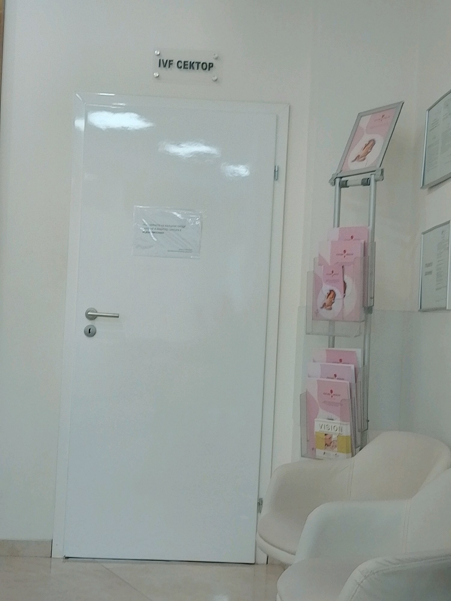The image shows a hallway in what appears to be a medical clinic or office. A white door with a silver handle and lock is on the left side of the frame. Above the door, a sign reads "IVF СЕКTOP," indicating an IVF department. A clear plastic sign with text is affixed to the door, partially obscured. To the right of the door, a literature stand displays several brochures, most of which are pink and feature images of babies or pregnant women. One brochure is labeled "VISION." In the foreground on the right, two white chairs are visible. The walls are a light, neutral color, and the floor is tiled in a beige or light brown color. The lighting appears to be artificial. The overall impression is one of a clean, professional, and modern waiting area or corridor within a healthcare facility. There are no people visible in the image, and no actions or interactions are depicted. This scene is located in Sofia, Bulgaria, as suggested by the Cyrillic text on the sign.