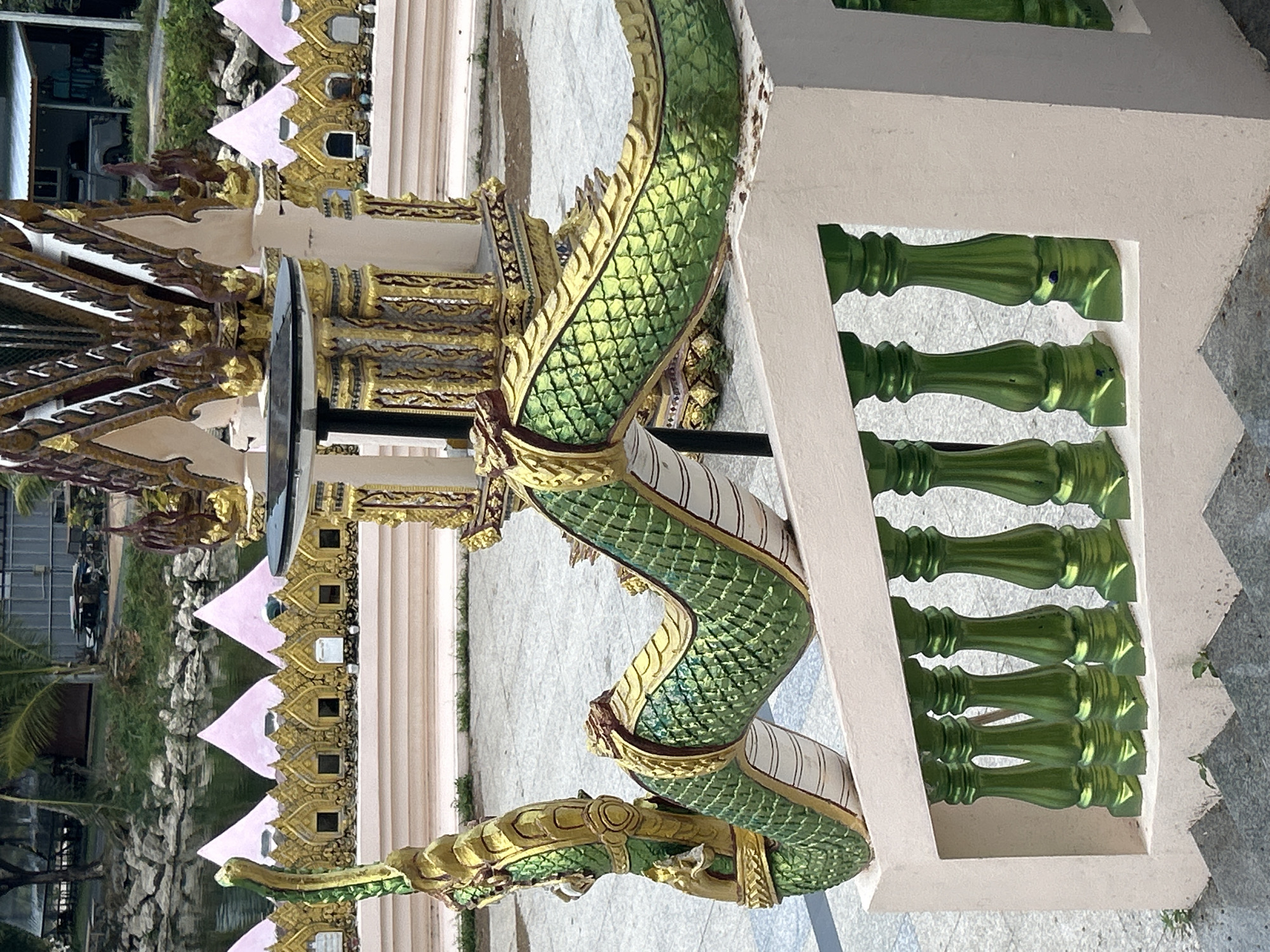 The image depicts a section of an ornate, possibly religious, structure in Ko Samui District, Thailand. In the foreground, a cream-colored railing with metallic green balusters lines a staircase. Partially visible on the railing is a decorative sculpture of a green, scaly serpent or dragon, its body curving and twisting. The serpent's scales are highly detailed, and it is adorned with gold accents. Behind the railing, a patterned walkway or plaza is visible. Further in the background, a series of golden, tiered architectural elements, reminiscent of a temple or shrine, rise upwards. A body of water, possibly a pond or canal, is situated beyond the structure, bordered by rocks and lush greenery. In the distance, a modern building with a white roof and a pickup truck parked in front suggests the presence of nearby habitation or facilities. The overall scene conveys a sense of cultural artistry and the integration of traditional architectural motifs with the natural and contemporary environment of the region.