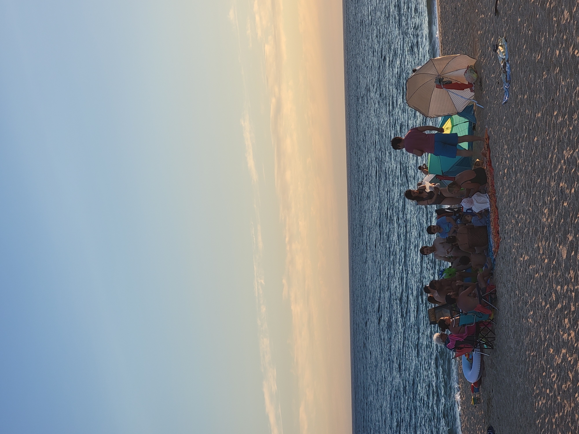 A group of people are gathered on a sandy beach in Quarteira, Portugal, near the ocean. The sky is a gradient of light blue with a band of pale orange and pink clouds near the horizon, indicating sunset or sunrise. The ocean is a deep blue with visible waves.
In the foreground, several people are sitting and standing around a small green tent and a large, striped beach umbrella. Some individuals are seated in beach chairs, while others are on blankets. One man, wearing a pink shirt and blue shorts, stands near the tent, facing away from the camera. A woman in a black swimsuit is standing next to him, holding a white object.
Further to the left, a group of people are seated on chairs and blankets. A white inflatable ring is on the sand to the left of this group. A collection of small buckets are visible near the inflatable ring. In the background, more people are visible near the water's edge. The sand appears textured and marked by footprints. A few towels are spread out on the sand in the mid-ground. The overall scene suggests a relaxed gathering on the beach during the late afternoon or early evening.