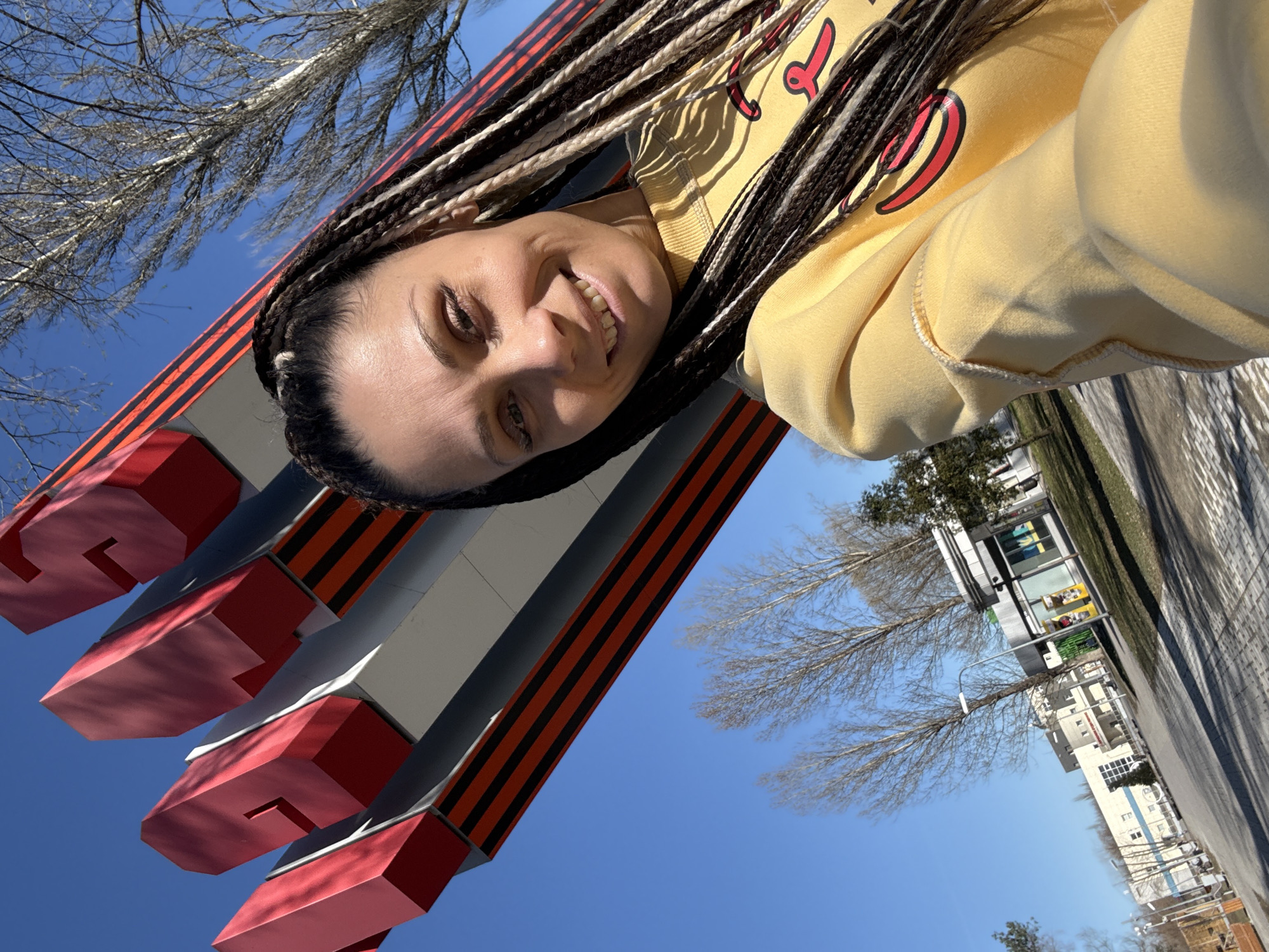 A woman with braided hair, wearing a yellow sweatshirt, is smiling at the camera. She is standing in front of a monument with red and gray blocks, the base of which is decorated with the Saint George ribbon (black and orange stripes), indicating it is a war memorial, possibly related to Victory Day celebrations in Russia. The location appears to be an urban area in the Rusanovo district of Moscow, Russia, with trees, buildings, and a street visible in the background. The sky is clear and blue.