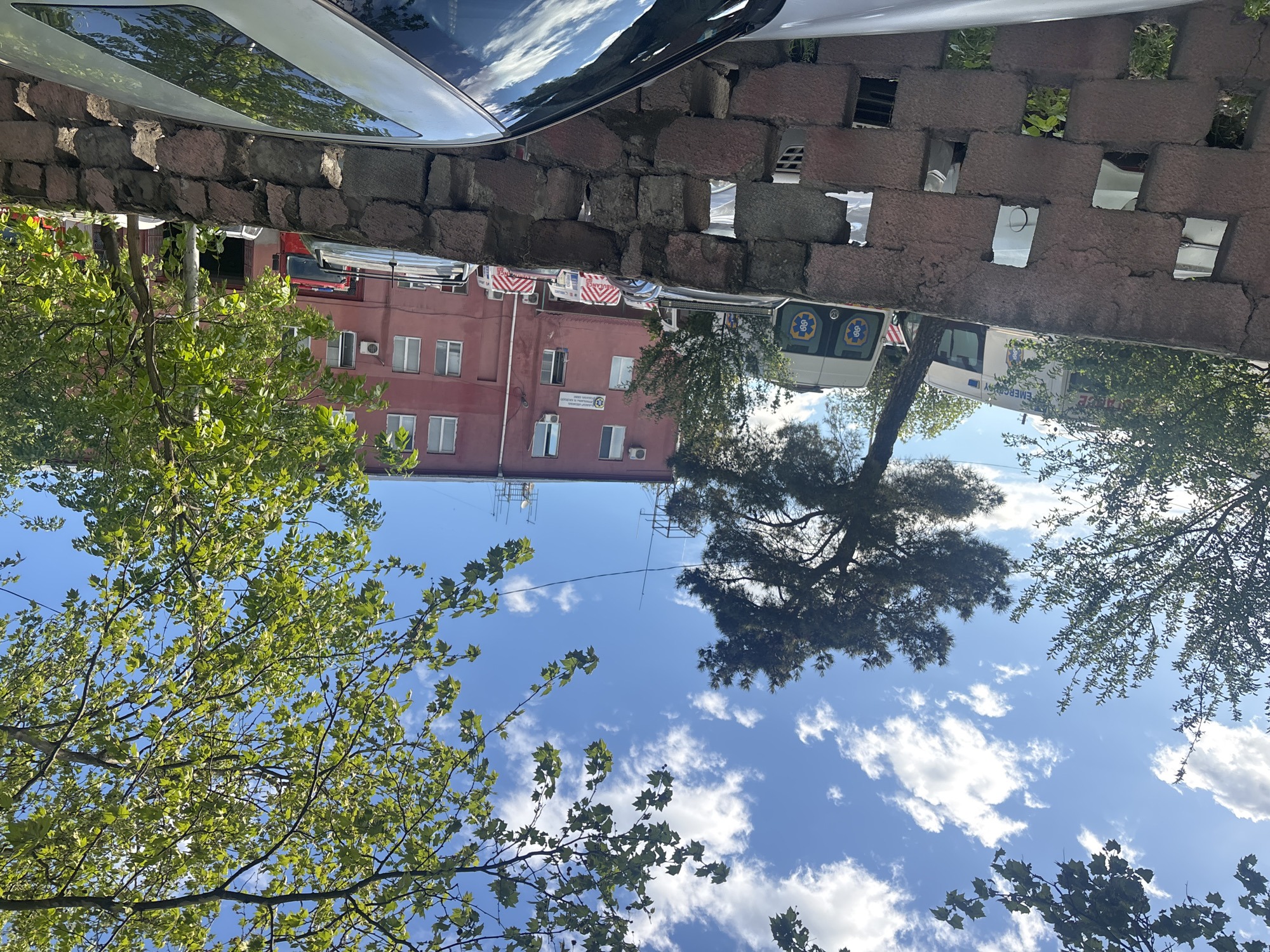 The image shows a view upwards towards a building with a brick wall and a car above it. The building is a salmon color with white-framed windows and several antennae on the roof. Two ambulances are parked near the building. The foreground includes a blue sky with scattered white clouds and green leafy tree branches. The location is Tbilisi, Georgia, based on the language on the ambulance and building signs.