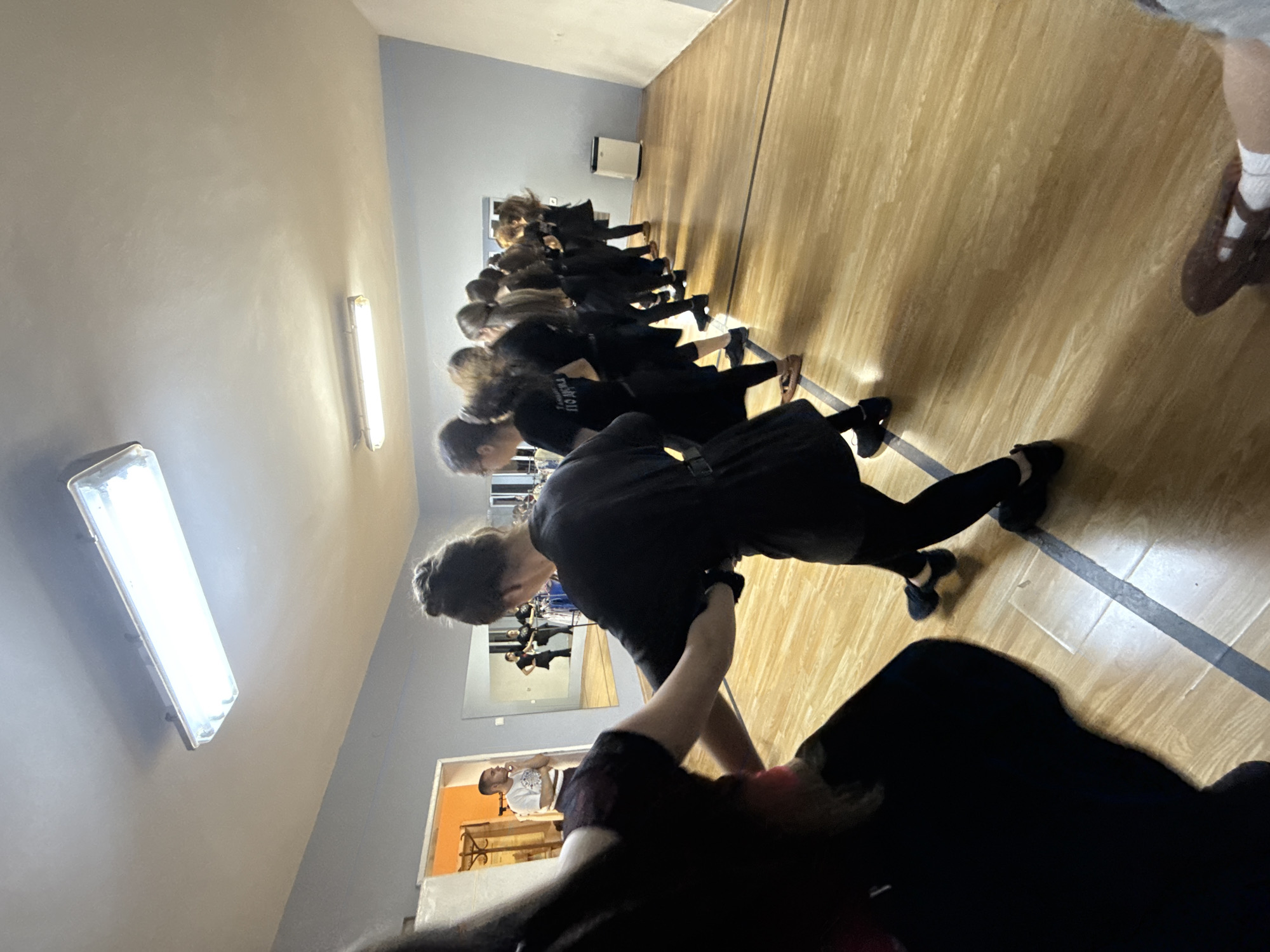 A group of approximately 10 people are lined up in a room with a wooden floor. Most of them are wearing black clothing. They appear to be participating in some kind of activity, possibly a dance or exercise class. A person is visible in a mirror and in an adjacent room. The location is possibly in Buhovo, Bulgaria.