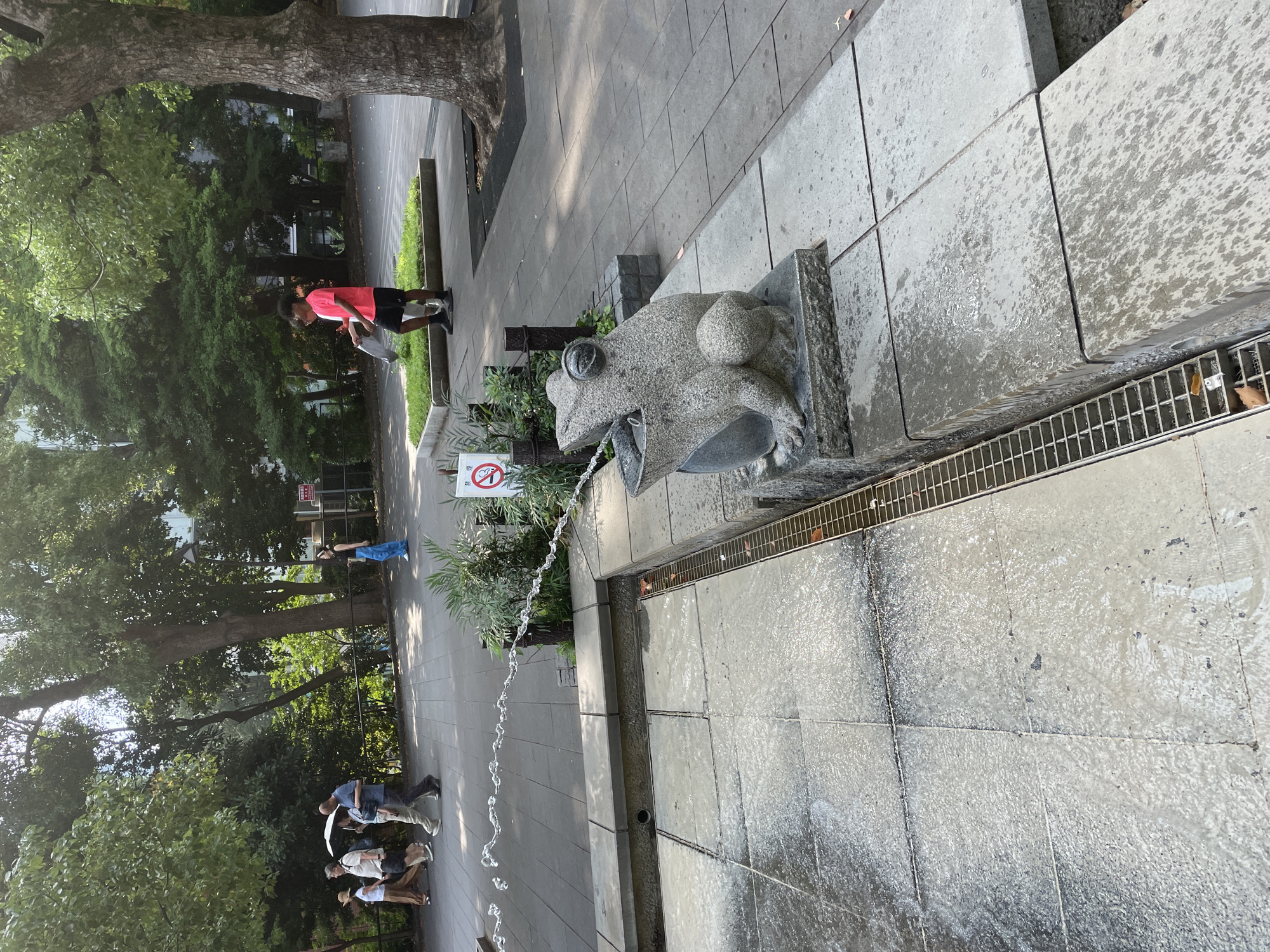 The image shows a stone frog fountain with water spraying from its mouth in Taito, Japan. A person in a pink shirt and black shorts is standing near the fountain, holding a plastic bag. Several people are walking in the background on a paved path lined with trees. The fountain is surrounded by stone tiles and a drainage grate. A "no smoking" sign is visible near the fountain. The scene appears to be in a park or public space with greenery and trees.