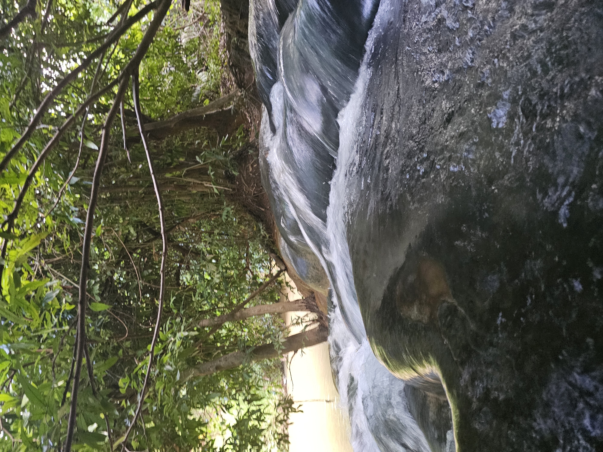 The image depicts a natural scene with a waterfall flowing over rocks, surrounded by lush green foliage. The water appears murky brown, suggesting it might be from a river or stream. Trees with dense green leaves line the upper part of the image, with some branches extending over the water. The rocks in the foreground are dark and appear wet from the cascading water. The setting is clearly outdoors, in a natural environment, consistent with the description of Tambon Khlong Thom Nuea, Thailand, known for its natural attractions. There are no people or man-made structures visible in the frame. The overall impression is one of a tranquil, undeveloped natural landscape.