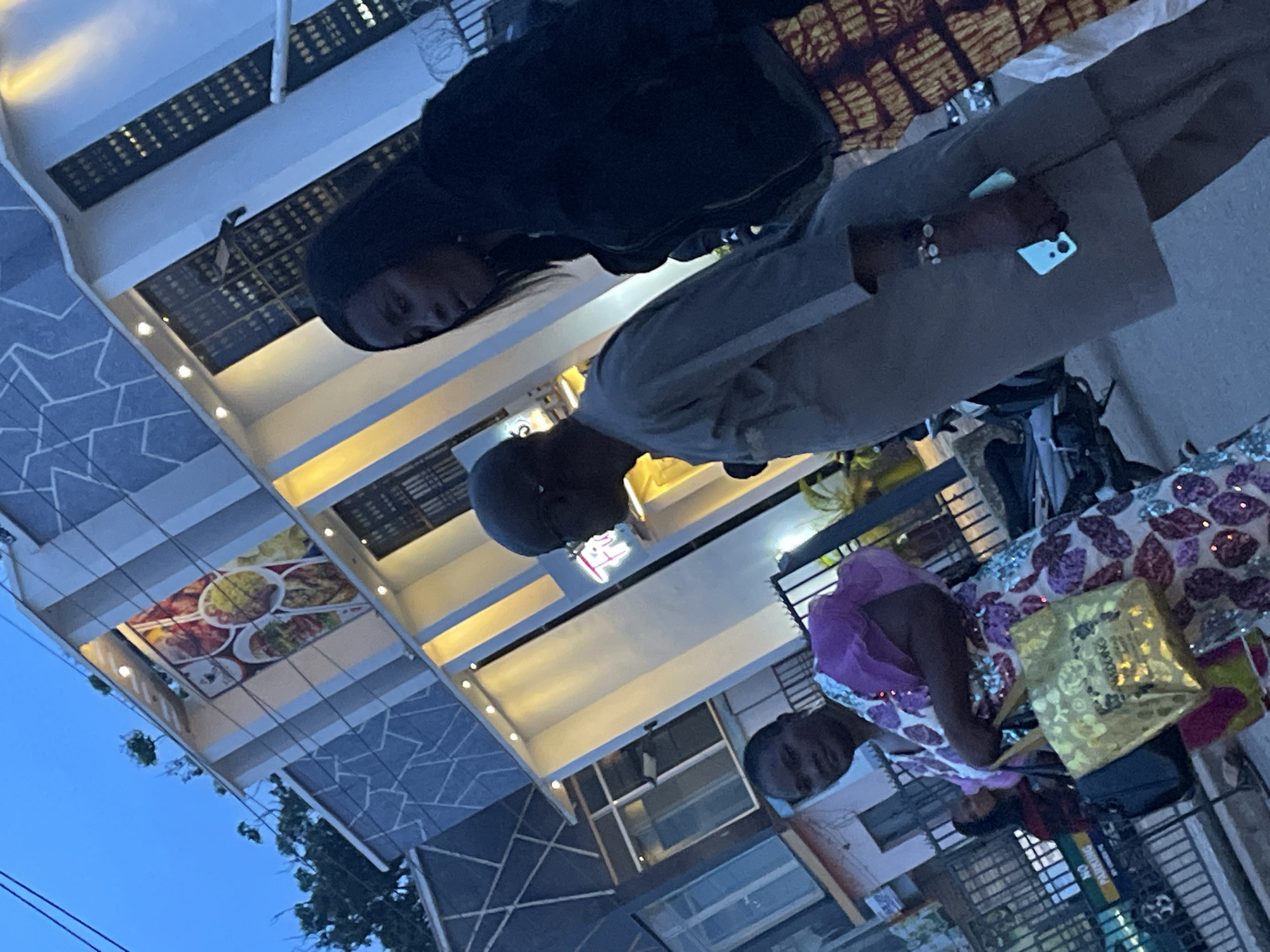 The image depicts a street scene in Jos, Nigeria, at dusk. In the foreground, three individuals are walking away from the camera. A woman on the left is wearing a vibrant, sequined dress and carrying two bags. Beside her, a man in a grey tunic is walking, and he is holding a light blue smartphone in his hand. To the right, another woman, partially obscured, is wearing a dark jacket and a patterned skirt. Behind them, a modern building with a patterned facade and illuminated vertical elements is visible. Trees and a bright blue sky with a few clouds form the background. The overall lighting suggests it is twilight.