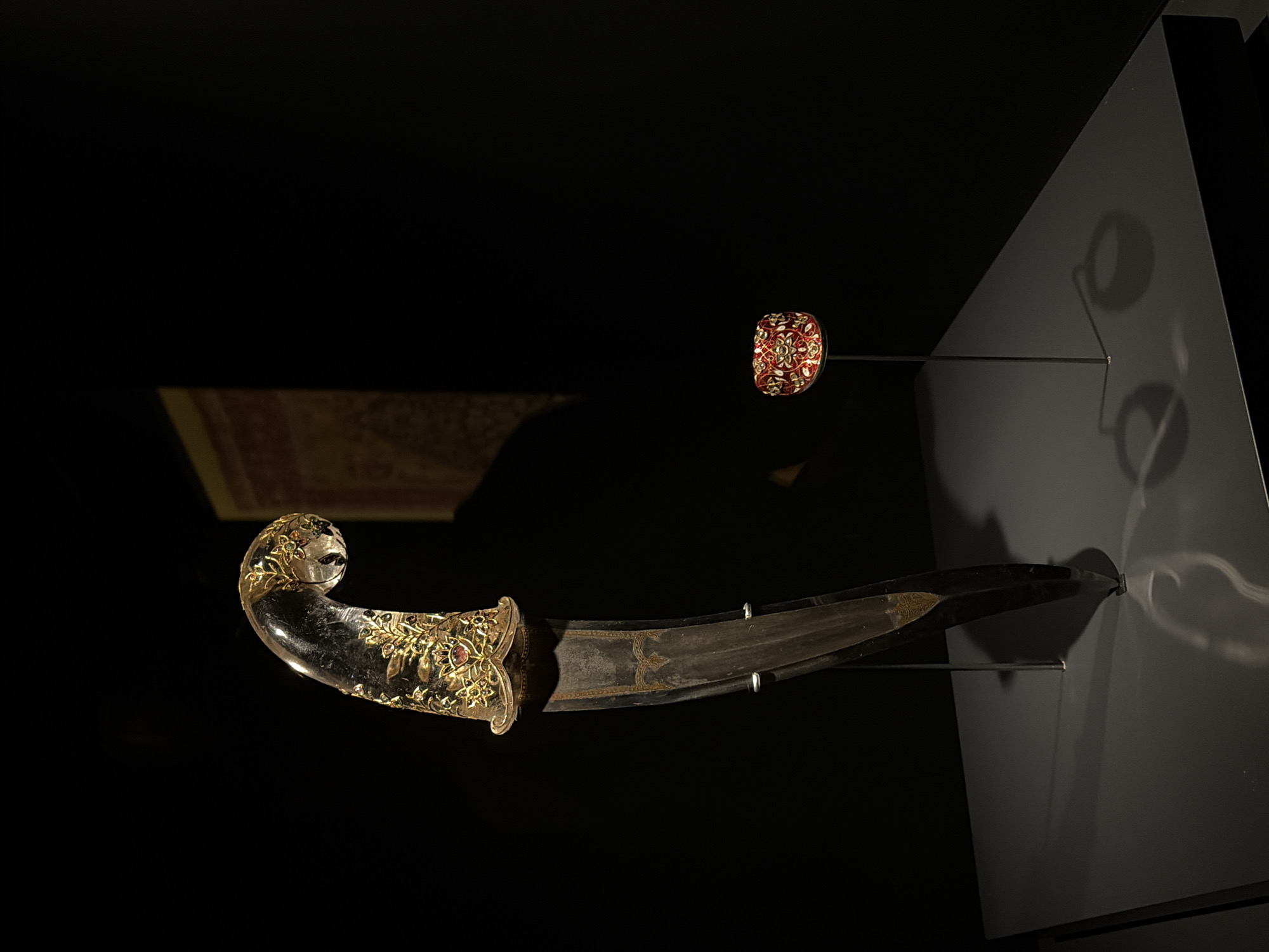 This is a medium shot of a display in a museum. The primary subject is a curved dagger with a transparent hilt encrusted with gold and precious stones. The dagger is positioned vertically, with its blade pointing downwards. To the right and slightly in front of the dagger, a small, ornate circular object, possibly a ring or a decorative cap, is displayed on a thin metal stand. Both objects are illuminated by spotlights, casting distinct shadows on the dark gray display surface. The background is almost entirely black, with a faint, out-of-focus glimpse of a tapestry or artwork with a red and gold pattern in the upper right corner. The overall presentation suggests a formal exhibition setting. The location is not determinable from the image.
