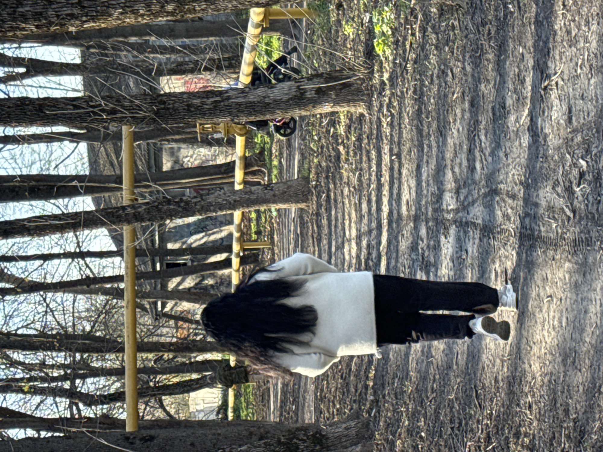 A person with long dark hair is walking on a dirt path in a park, wearing black pants, white shoes, and a light-colored jacket. The path is surrounded by trees with bare branches. A yellow metal structure is visible on the left, possibly playground equipment. The ground is covered with leaves and debris. The scene appears to be set in Rostov-on-Don, Russia.
