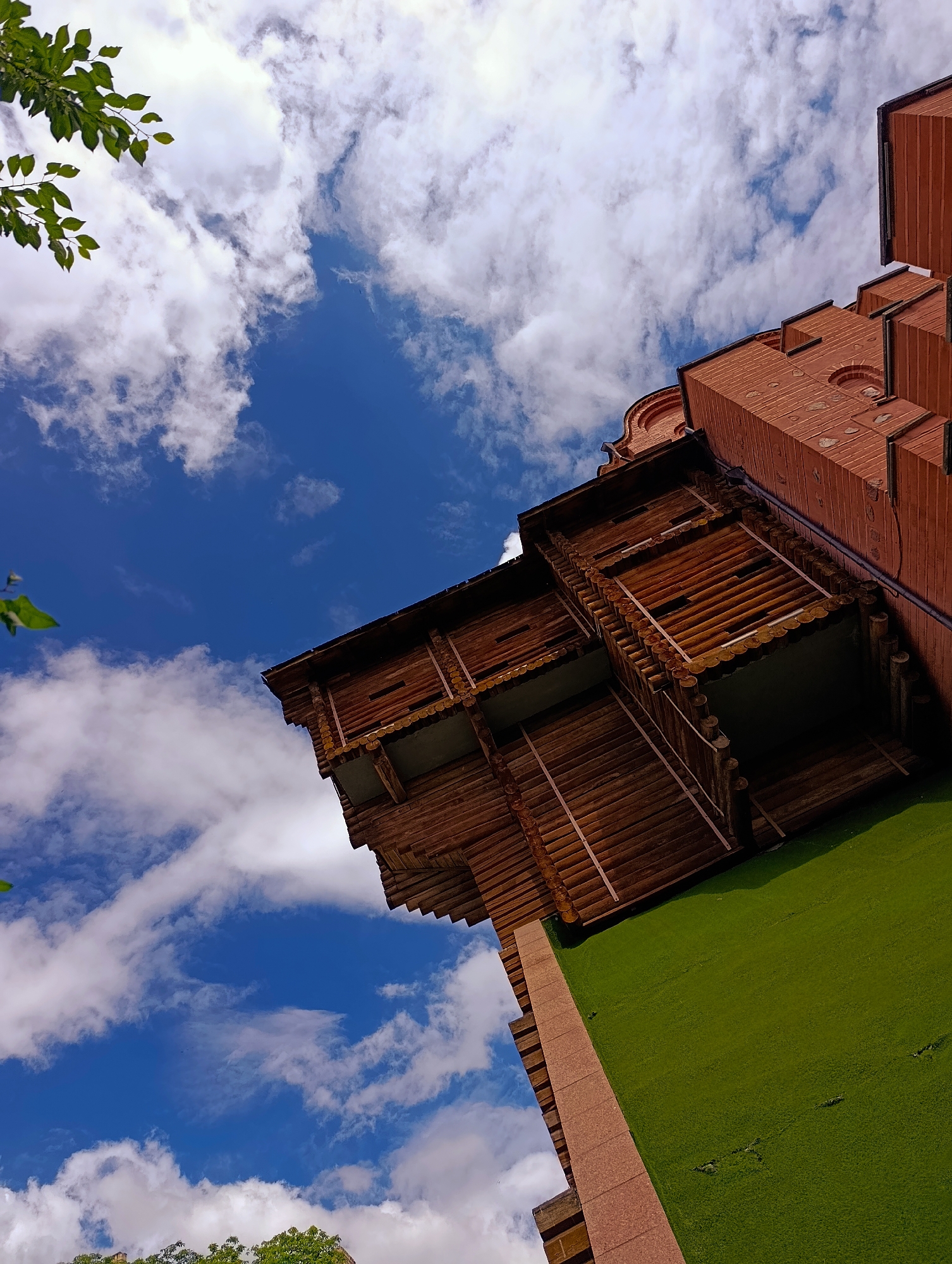 The image shows a low-angle shot of a modern building in Kyiv, Ukraine, with a prominent blue sky filled with white clouds. The building appears to be a multi-story structure with a unique architectural design. The lower portion of the building features a bright green surface, possibly artificial turf or a similarly colored facade. Above this green section, there is a wooden structure with multiple levels and balconies, constructed from logs. The upper part of the building is made of red brick, with decorative elements and visible mortar lines. In the top left corner of the image, a branch with green leaves extends into the frame, suggesting the presence of trees nearby. The overall scene depicts an urban setting with a blend of natural and architectural elements under a bright, cloudy sky.