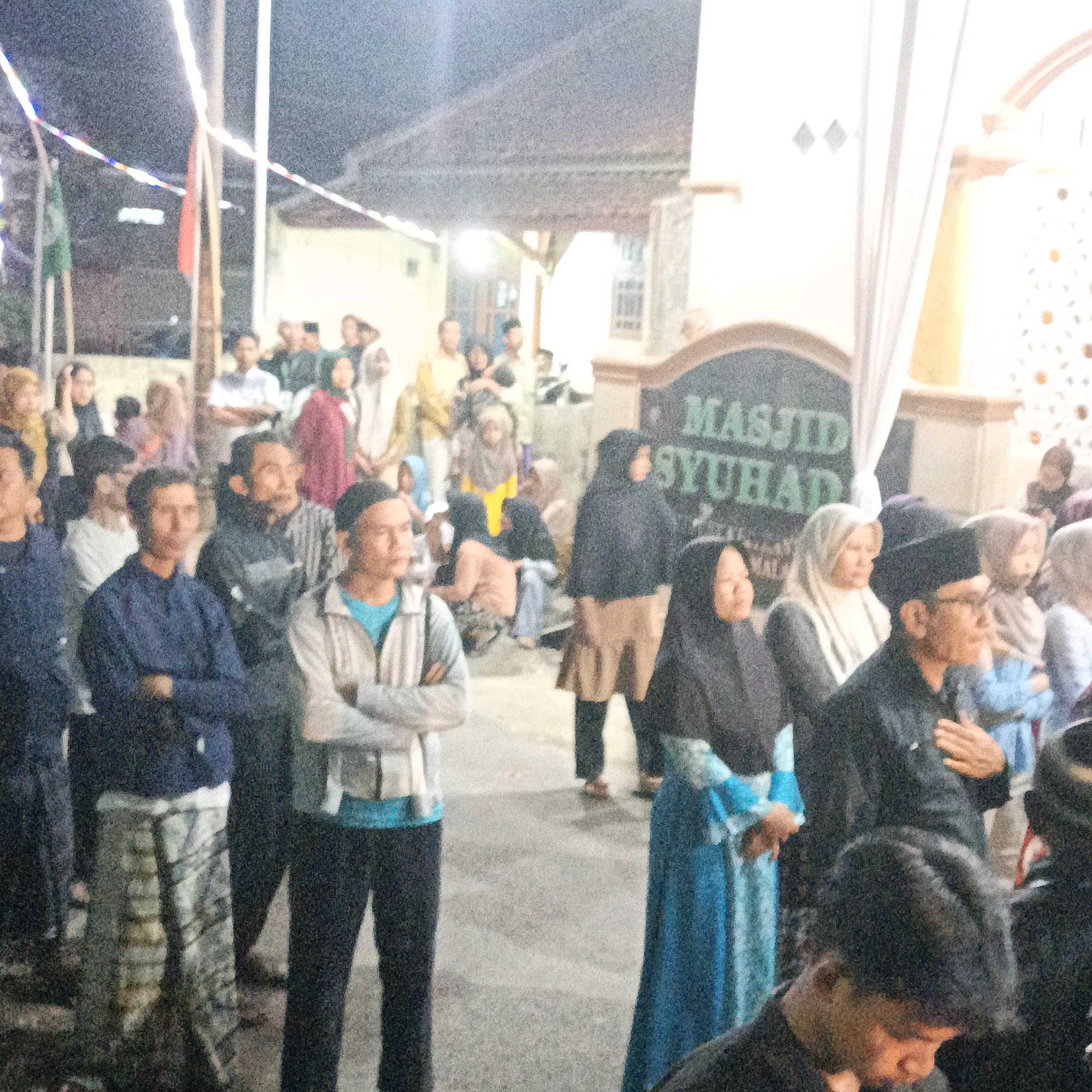 The image depicts an outdoor gathering of people, likely in the city of Kalitorong, Indonesia, possibly near a mosque based on the visible sign "MASJID SYUHAD". The lighting suggests it is evening or nighttime, with artificial lights illuminating the scene. A diverse group of individuals, predominantly men and women, are present, many wearing traditional Islamic attire such as headscarves and kufis. Some people are standing, while others are seated. There is a sense of communal activity, with individuals interacting and observing. In the foreground, several men are visible, some with their arms crossed. A large sign, partially obscured, identifies the location as a mosque. The environment appears to be paved, with some greenery and buildings in the background. The overall atmosphere suggests a religious or social event.