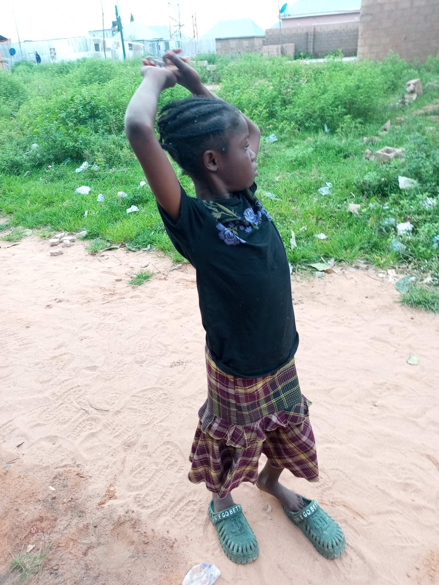 A young girl with braided hair stands on sandy ground, facing right, with her arms raised above her head. She wears a black t-shirt with a floral design around the neckline and a plaid skirt. Her green sandals have the words "GO BETTER" imprinted on them. The background shows a grassy area with scattered debris and buildings in the distance, indicative of a semi-developed urban environment in Tempure, Nigeria. The ground is marked with numerous footprints.