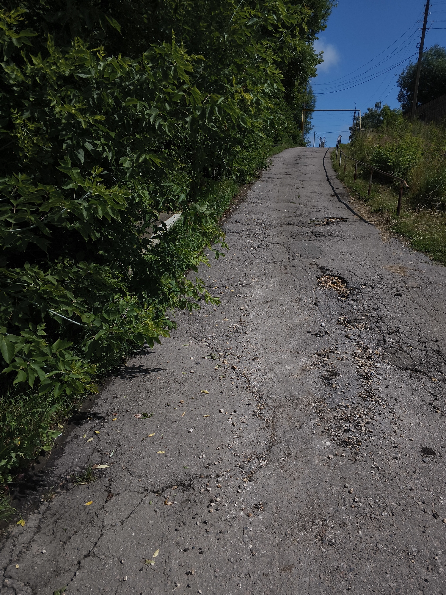 The image shows a steep, cracked asphalt road winding uphill. Dense green foliage lines the left side of the road, with grass and wild plants growing on the right. A rusty metal handrail runs along the right side of the path. In the distance, utility poles with wires and a yellow horizontal pipe structure are visible against a bright blue sky with a few white clouds. The road surface is uneven, with several potholes filled with loose gravel. The scene appears to be in a somewhat neglected, rural or suburban area, possibly a back road or a path leading to a more remote location. The lighting suggests it is daytime, with the sun casting shadows from the vegetation. No people are present in the image.