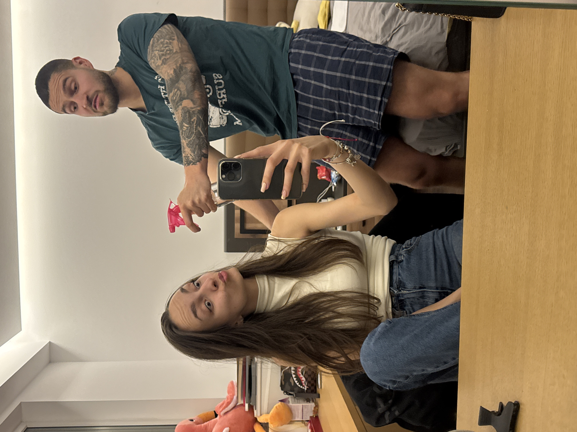 A man and a woman are taking a selfie in a bedroom. The woman, facing the camera, is wearing a white tank top and blue jeans, and has long brown hair. She is holding a black iPhone. The man, behind and to the side of her, is wearing a green t-shirt, blue plaid shorts, and has tattoos on his left arm. He is holding a pink spray bottle. The background shows a bed with gray bedding, a wooden headboard, and a light-colored wall. There is a wooden desk with personal items. The location is likely an apartment in Sofia, Bulgaria, based on the modern interior design and common building styles.