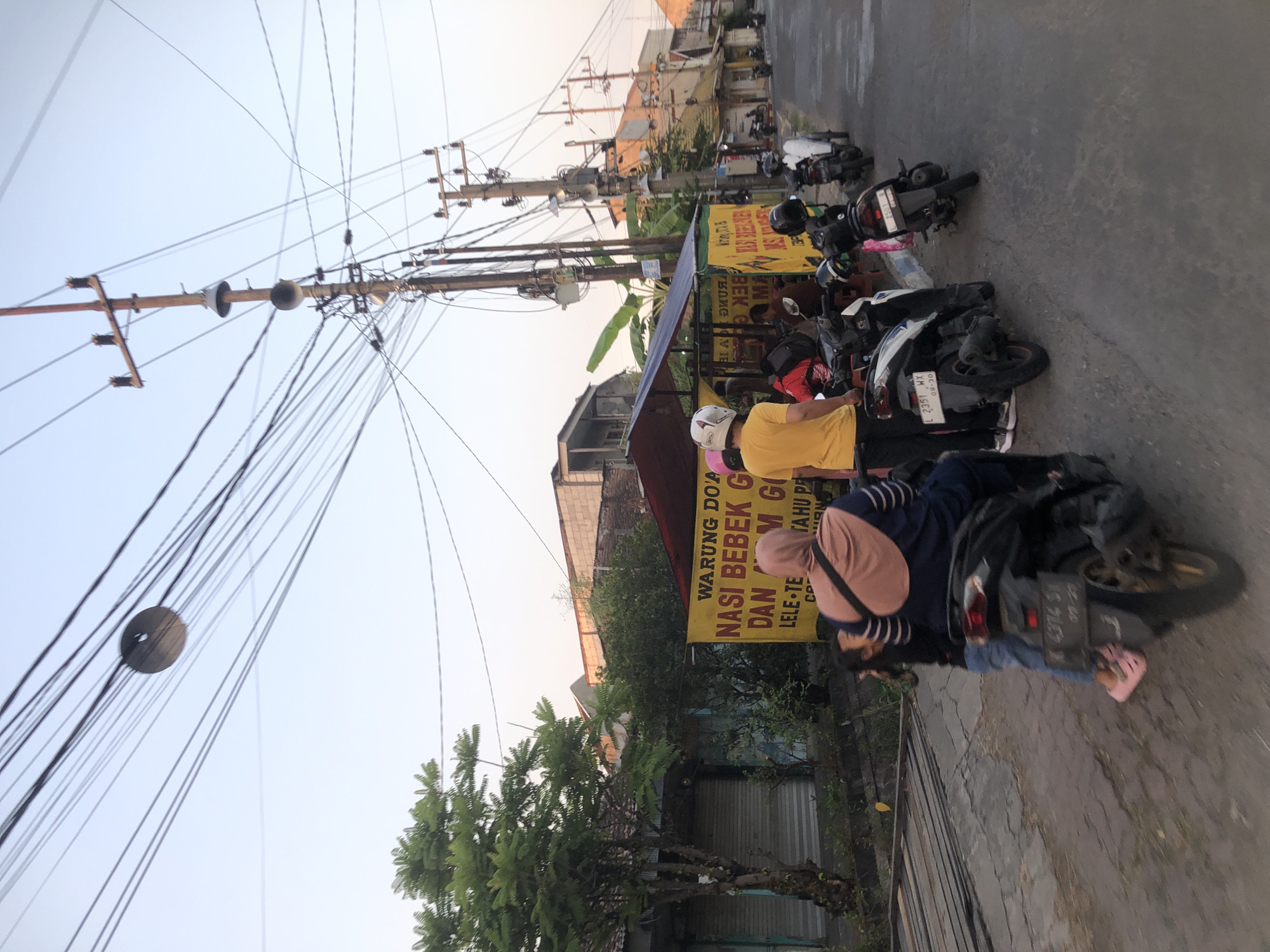This image captures a street scene in Sidoarjo, Indonesia, likely in a residential or commercial area given the presence of shops and overhead utility lines. A food stall with a yellow banner, displaying "WARUNG DOA NASI BEBEK GORENG M G", is a focal point. Several people are gathered around the stall, with some on motorcycles. Two women and a child are on a dark-colored scooter in the foreground, with the child facing away from the camera. Another individual in a yellow shirt and black pants stands near a white and blue scooter. Several other motorcycles are parked or in use on the street, which appears to be paved. Overhead, a complex network of electrical wires crisscrosses the sky, supported by wooden utility poles. The lighting suggests it is either early morning or late afternoon. The environment includes trees, buildings with varying architectural styles, and what appear to be closed storefronts on the left.