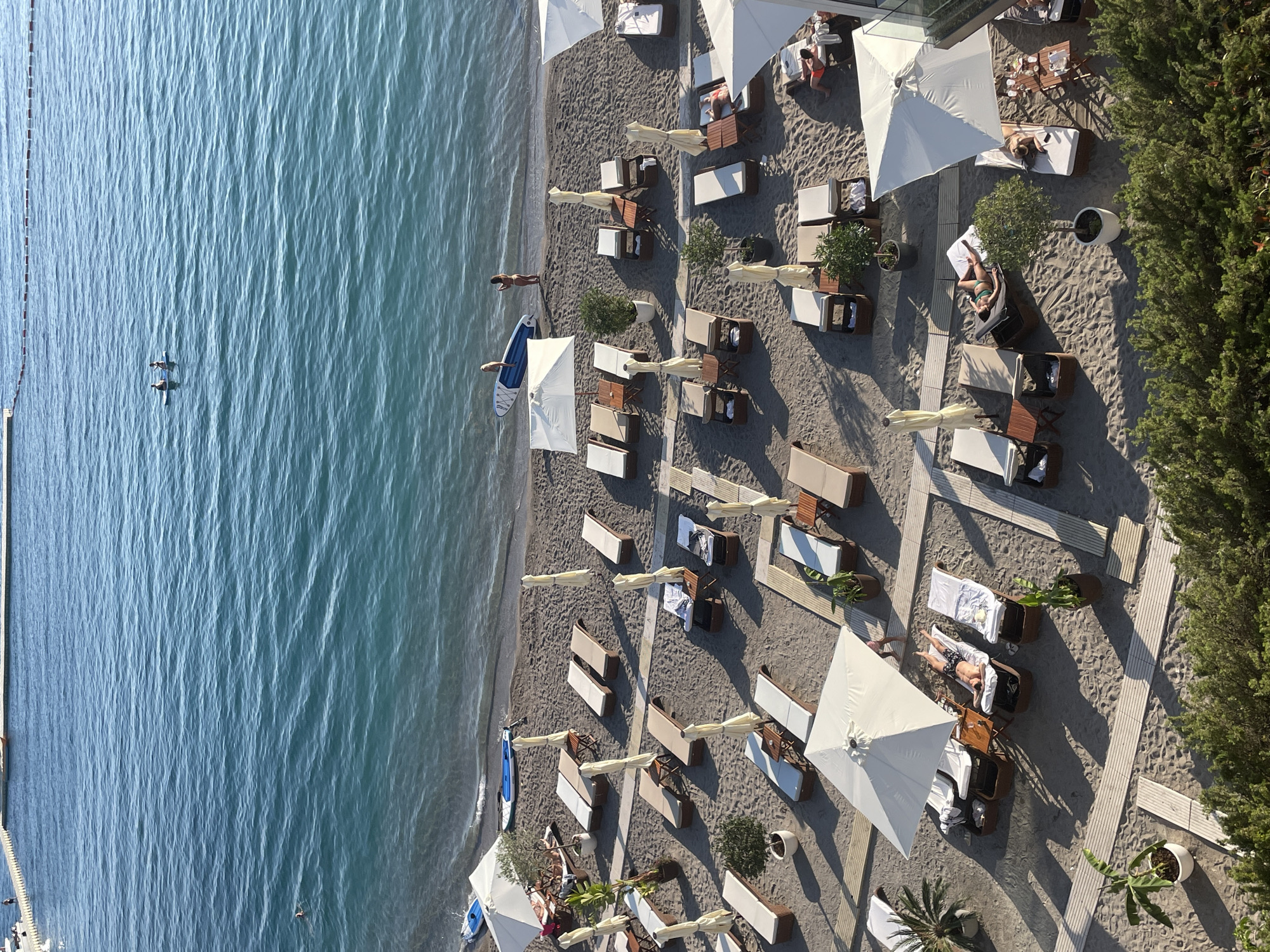 Aerial view of a beach in Budva, Montenegro, with a shoreline featuring turquoise water on the left. A pier extends into the water on the far left. Two kayakers paddle on the water. The sandy beach is arranged with rows of lounge chairs and umbrellas. People are relaxing on the lounge chairs. Bushes and trees border the edge of the beach. The scene conveys a tranquil, sunny atmosphere.