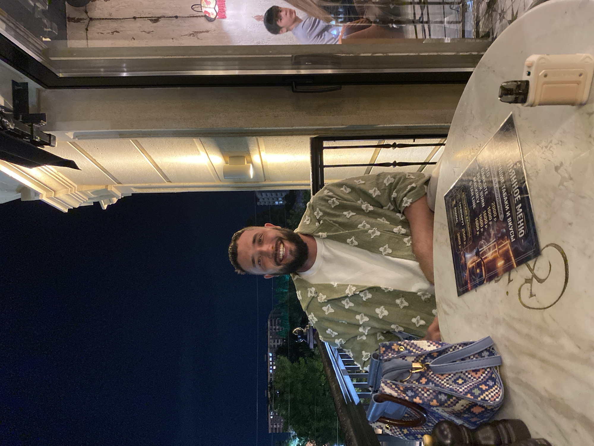 A man with a beard and short hair is sitting at a round table on a balcony at night. He is wearing a white t-shirt and a light green shirt with a white floral pattern. He is smiling broadly at the camera. In front of him, on the table, are a menu and a vape device. To his left, a patterned blue and white handbag is placed on the table. In the background, through a glass door, another person, a young male, is visible sitting at a table inside. The balcony has a railing, and behind it, there are trees and buildings. The building facade on the right side of the image is lit by warm lights. The scene is located in Derbent, Russia.