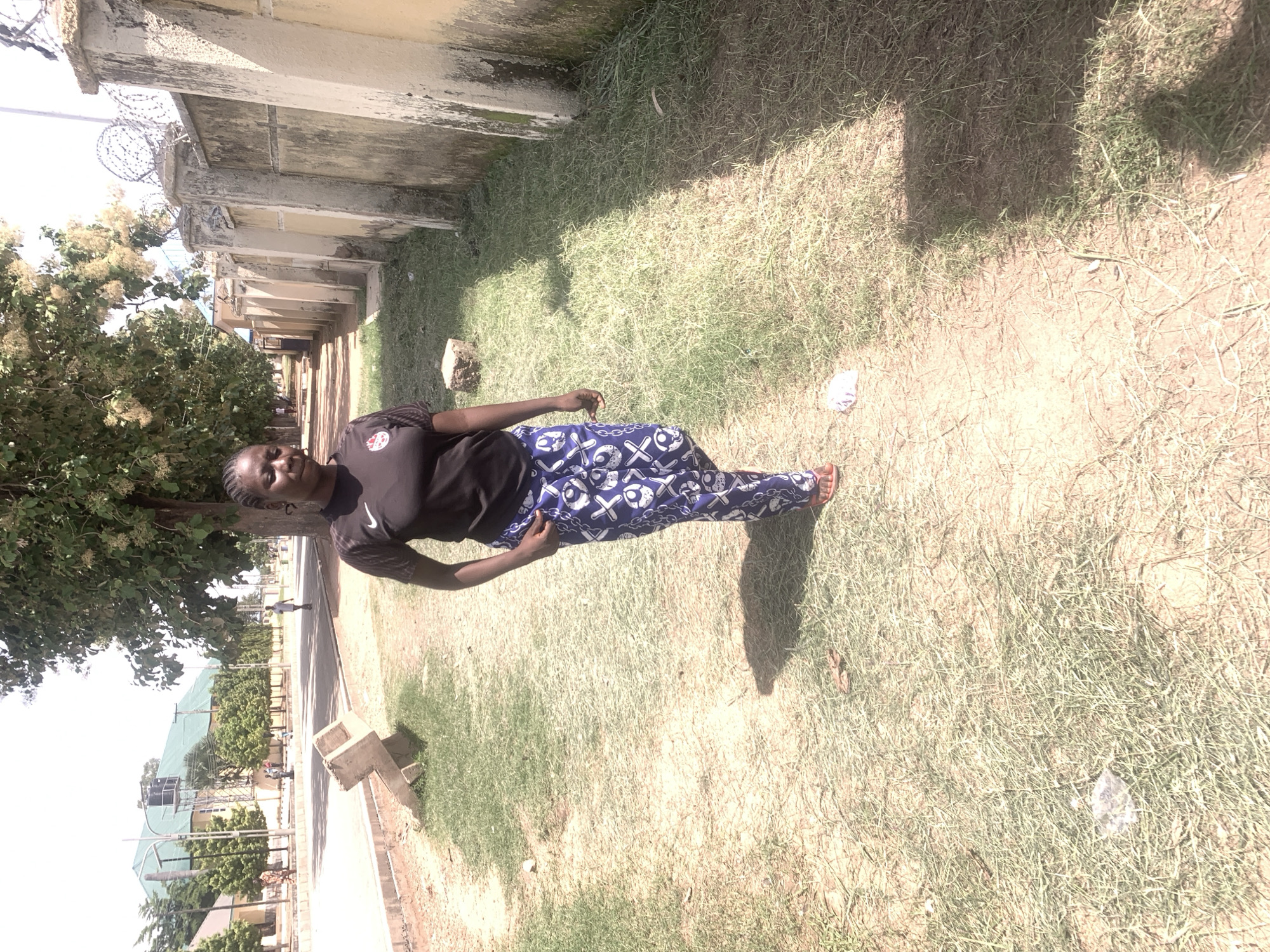 The media file depicts a young Black woman standing outdoors on a patch of dry grass, with a wall and barbed wire in the background. She is wearing a black t-shirt and blue patterned pants. The woman is looking towards the camera with a slight smile. The scene appears to be a sunny day, with shadows cast on the grass, suggesting it is daytime. The setting shows a dry, unkempt grassy area next to a weathered wall, implying an outdoor urban or semi-urban environment. In the distance, there are buildings with green roofs and trees, and a road with a few people visible. The overall mood seems relaxed and casual. There is no visible text in the media file. The image orientation is sideways.