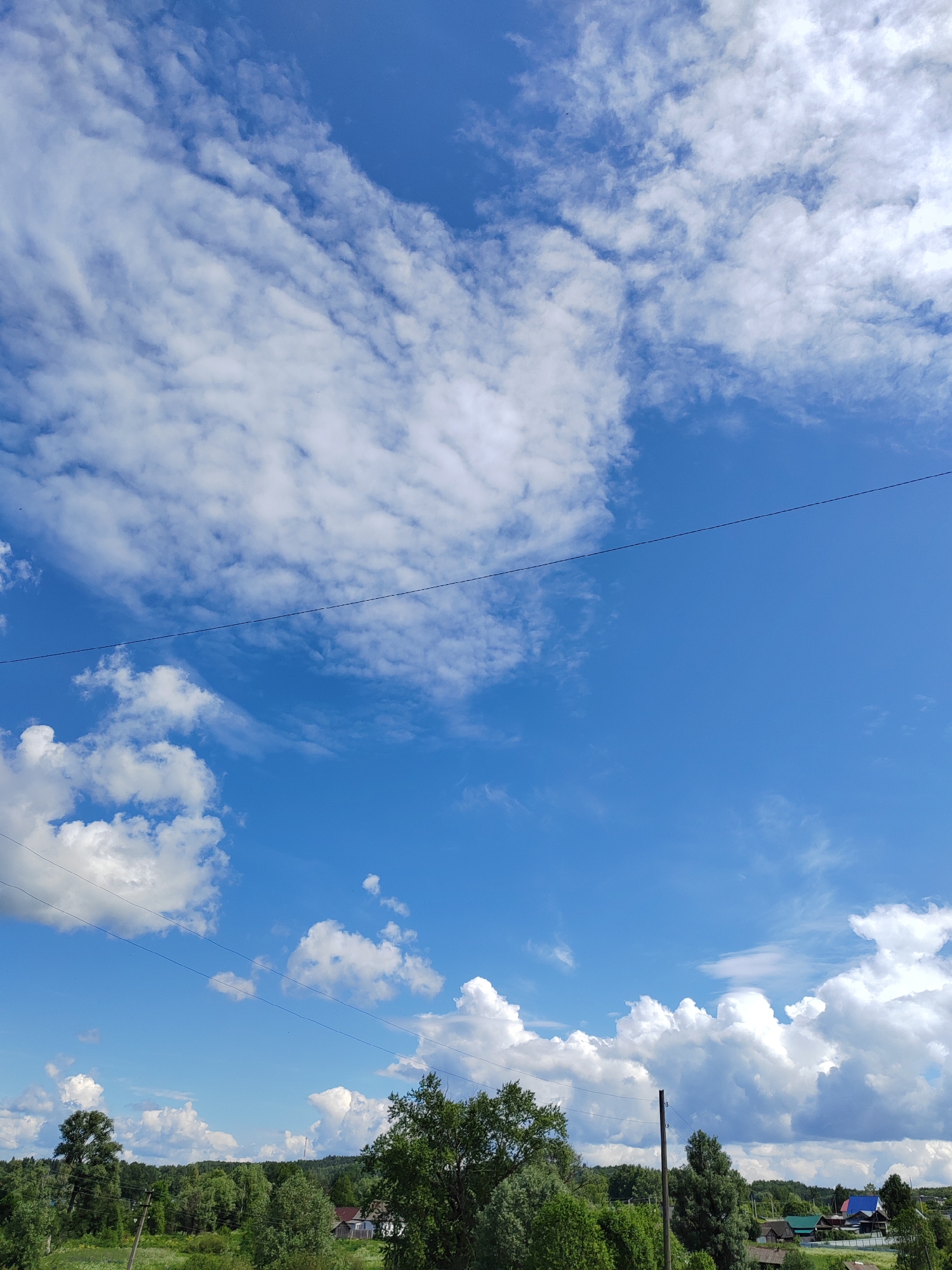 The image captures a broad rural landscape under a clear, bright blue sky with extensive white cumulus and cirrus clouds. The upper left portion of the sky is dense with patchy, diffuse white clouds, transitioning to a clearer blue expanse towards the right and lower center. A thin, dark utility wire spans horizontally across the upper middle of the frame. In the lower half, a dense line of green deciduous trees and foliage dominates the mid-ground, extending from left to right, with varying shades of green indicating diverse vegetation. Several utility poles with wires are visible among the trees, one prominently on the left and another on the right. In the distance, nestled within the treeline, are several residential structures with distinctly colored roofs, including blue, green, and red, primarily on the right side. The overall scene depicts a sunny day in a verdant, semi-developed countryside environment, specifically in the city of Unknown, Unknown.