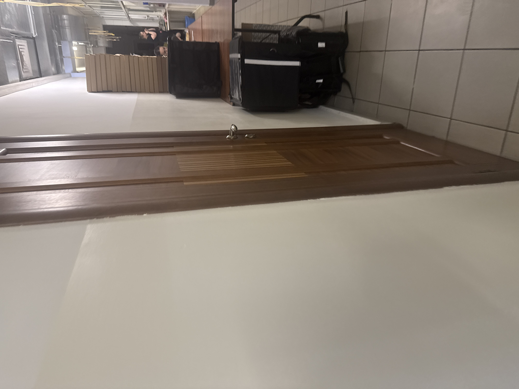 The image shows a narrow hallway with a wooden door on the left and a wall on the right. On the right side of the hallway, there is a table with two large black insulated bags on it. The bags appear to be delivery bags. Behind the table, there are some people in the background, suggesting a public or commercial space. The floor is tiled. The lighting is artificial and appears to be from overhead fixtures. The overall scene suggests a delivery service or a place where food is prepared and distributed. This location is not identifiable as Birobidzhan, Russia, or any other specific city.