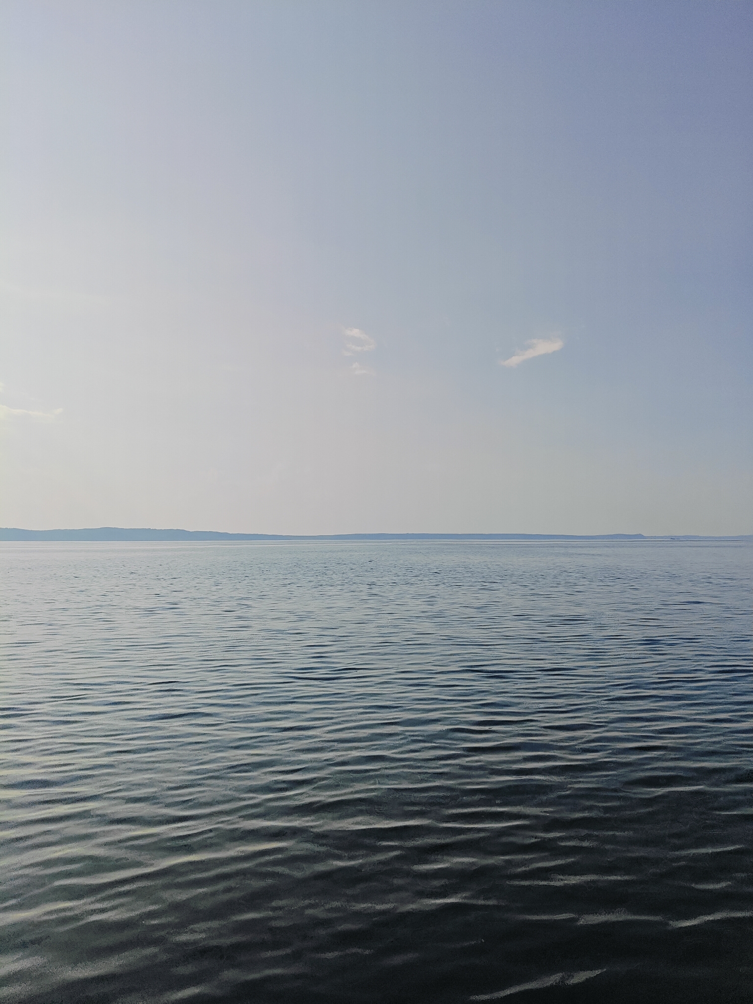 The image depicts a serene expanse of water under a pale, overcast sky. The water surface is rippled, creating a pattern of light and shadow that extends towards a distant, flat horizon. A faint, undulating landmass is visible along the horizon line. There are no people, animals, structures, or any man-made objects in the frame. The scene appears to be a natural landscape, likely a large lake or reservoir, given the scale and flatness of the water and the distant shoreline. This imagery is consistent with views of the Kama Reservoir, which is a significant geographical feature adjacent to the city of Chaykovsky, Russia. The overall impression is one of tranquility and vastness.