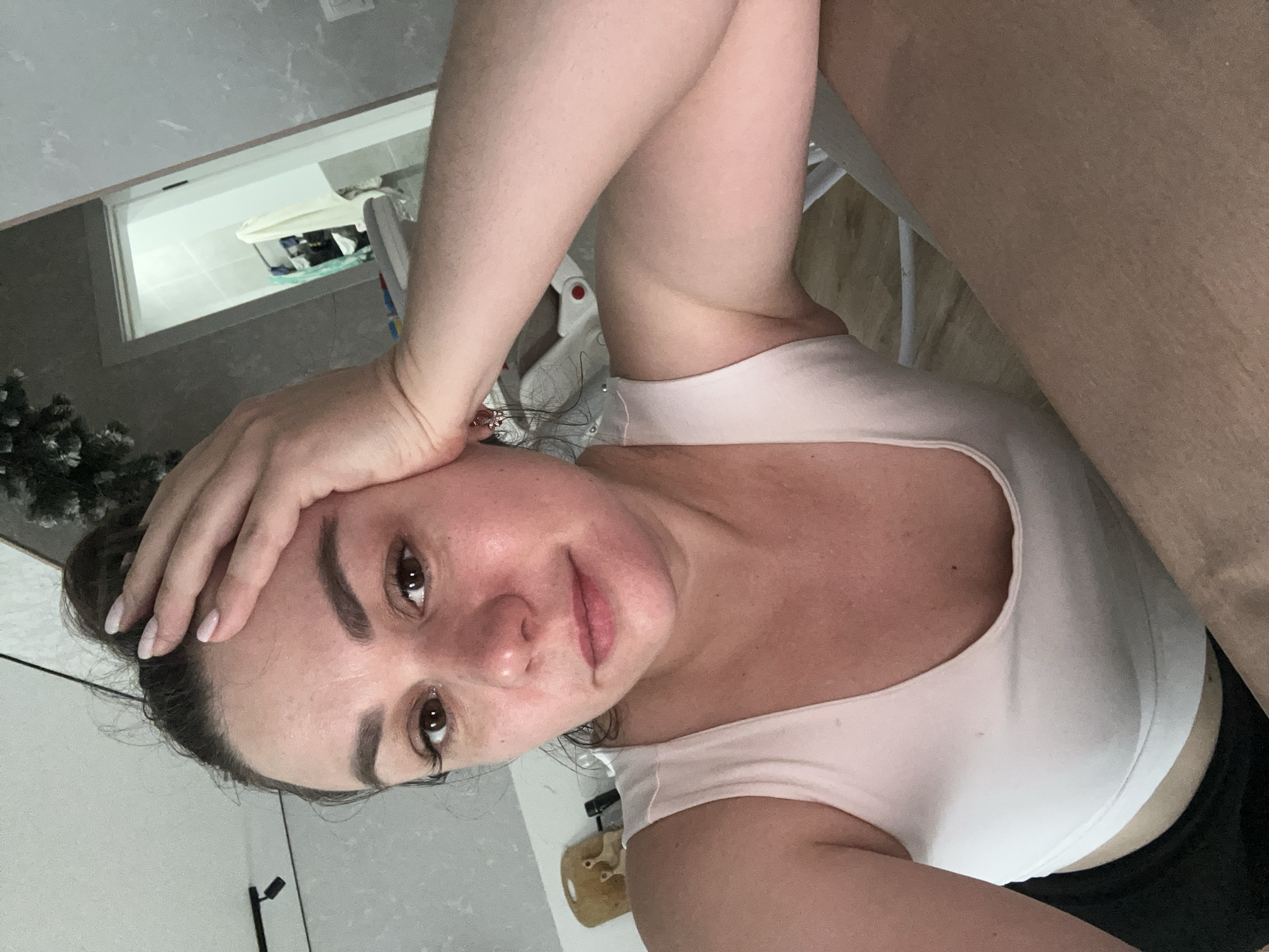 A medium-shot selfie captures an adult female indoors. She gazes directly at the camera with a neutral to slightly weary expression. Her dark hair is styled in a messy bun on top of her head, and her left hand, adorned with light pink polished nails, rests on her forehead. She wears a light pink or off-white sleeveless top and visible black bottoms. Small gold-toned earrings are visible in her right ear. Her skin exhibits some natural redness on the cheeks and nose, and she has well-defined eyebrows and prominent eyelashes.
The subject is positioned in what appears to be a residential room with light gray, textured walls and a white ceiling featuring a black track light fixture on the left. A decorative element, possibly a frosted artificial tree or branches, is partially visible in the upper right background. Behind her left shoulder, a light-colored plush toy is discernable on a shelf. To her right, a portion of a white object with a red button, possibly a high chair, is visible. The lower right foreground shows a light brown surface, likely a table or desk, on which her right arm and a portion of her leg rest.
On the right side of the image, an open white doorway leads into another room with light-colored tiled walls, strongly suggesting a bathroom. Inside this room, a white towel and other fabric items are hanging on a rack. The specific location within Chaikovsky, Russia, is not discernible from the visual information provided in the image.