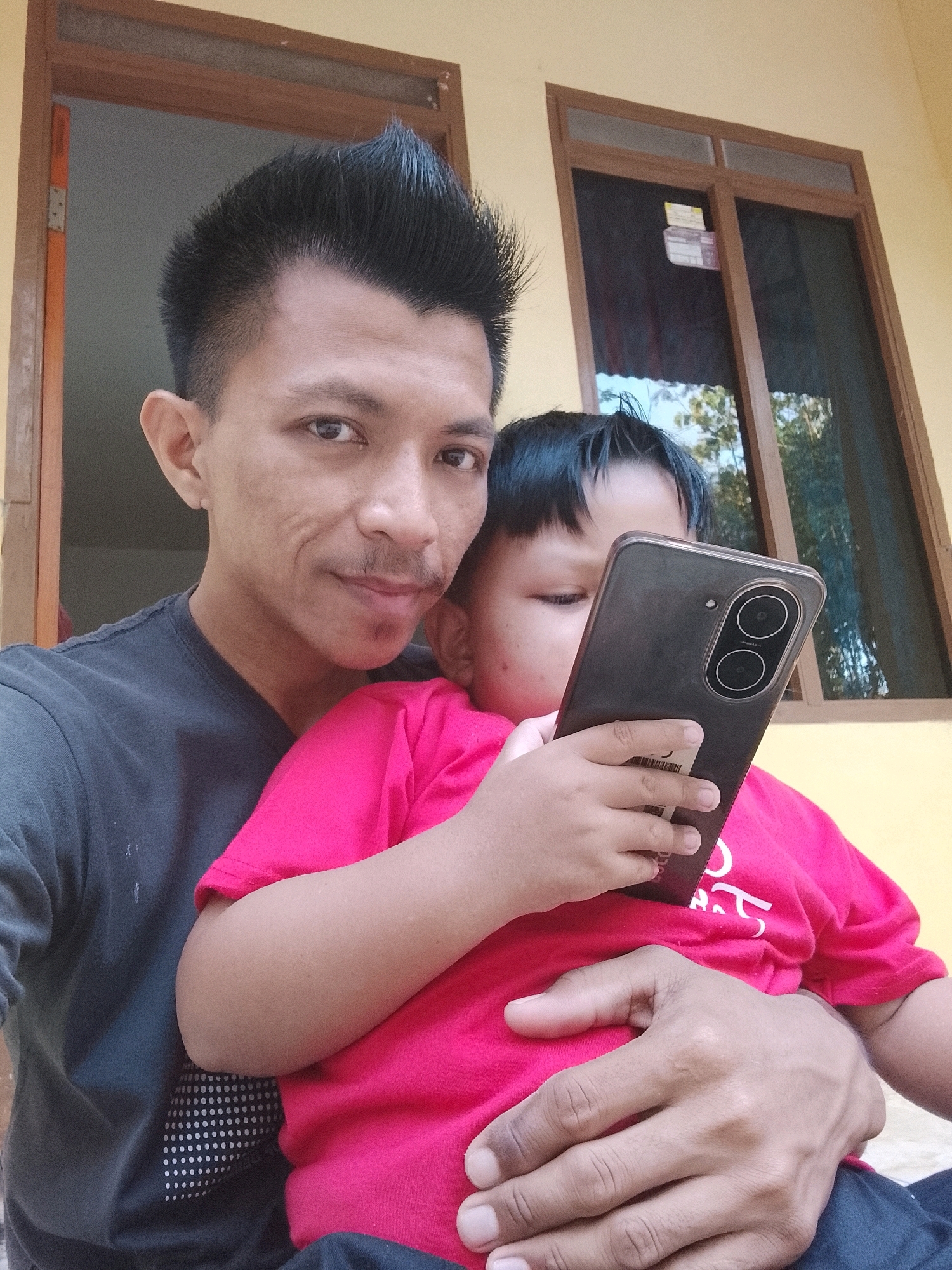 An outdoor selfie taken in Kertahayu, Indonesia, features a man holding a young child while they both look at a smartphone. The man, with dark, spiky hair and a mustache, wears a dark gray t-shirt. The child, wearing a pink t-shirt, is seated in the man's lap, holding the phone. The background includes a wooden door frame and a window with reflections of trees and sky, set against a yellow wall.