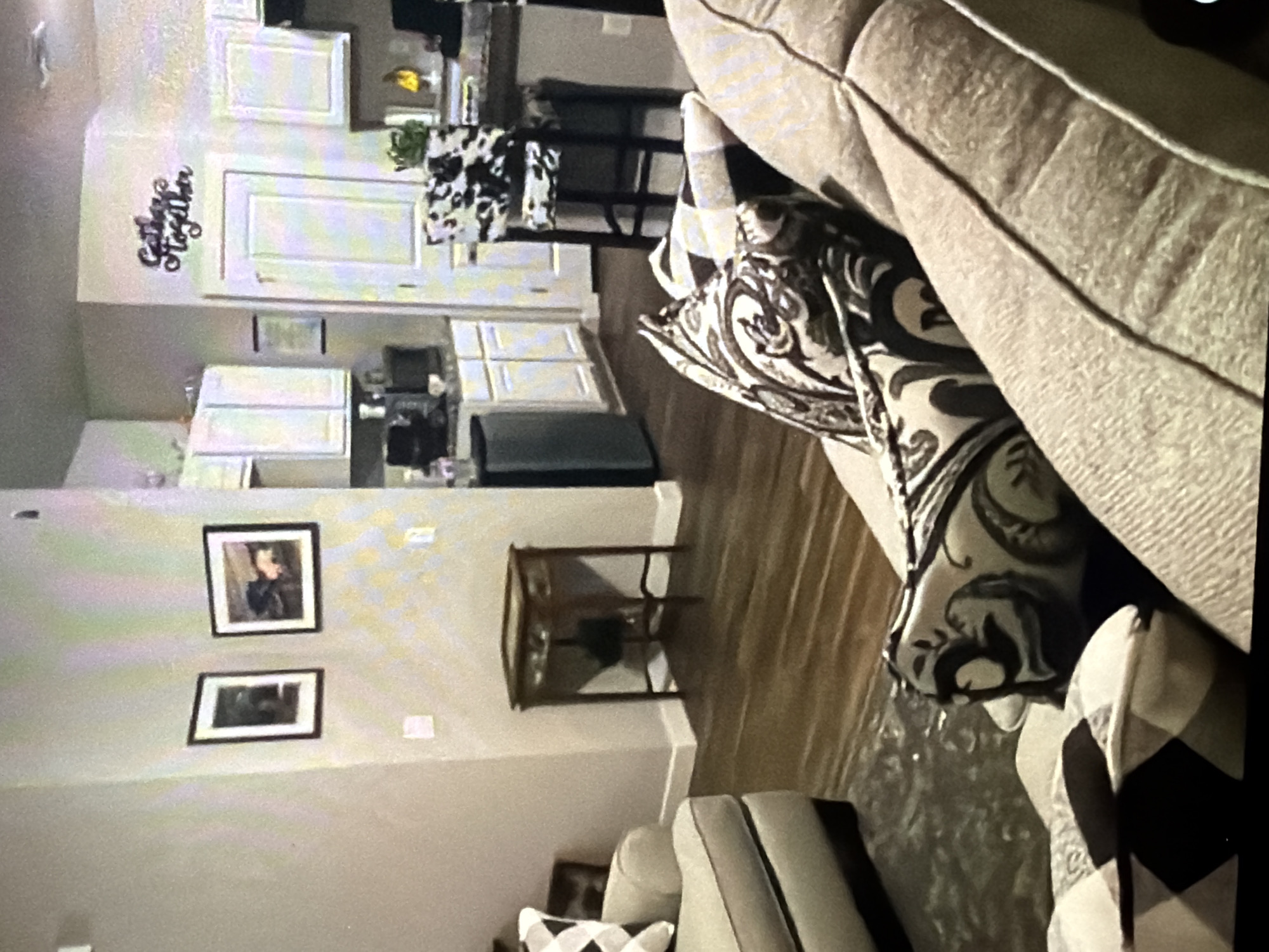 The image depicts a living room and kitchen area in a residential setting. In the foreground, a light-colored couch is visible with patterned throw pillows. To the left, another couch or chair with a patterned pillow is partially shown. A wooden accent table with drawers stands in the middle ground, near a wall with two framed pictures. The background reveals a kitchen with white cabinets, a refrigerator, and a counter area with a stool featuring a cowhide-patterned seat. A decorative sign with the text "Gather Together" is mounted on the wall above a closed door. The flooring appears to be wood-laminate throughout the space.