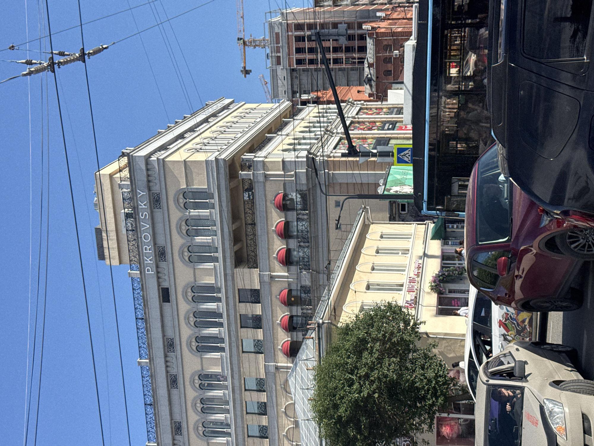 The image shows a street scene in Yekaterinburg, Russia, on a clear, sunny day. In the foreground, several cars are stopped in traffic, including a light-colored sedan on the left, a maroon SUV in the center, and a black SUV on the right. The light-colored sedan has some decals on its hood. Behind the cars, a large, multi-story building with classical architecture dominates the background. The building is light yellow with ornate detailing, including arched windows and balconies. The word "PKROVSKY" is visible in large letters on the facade of this building. To the right of this building, a modern building is under construction, with a crane visible at the top. A large, dark-colored bus is also present in the traffic, with reflections of people visible in its windows. A large green tree is situated to the left of the buildings. Overhead, power lines crisscross the sky. Several pedestrians are visible on the sidewalks and near the buildings.