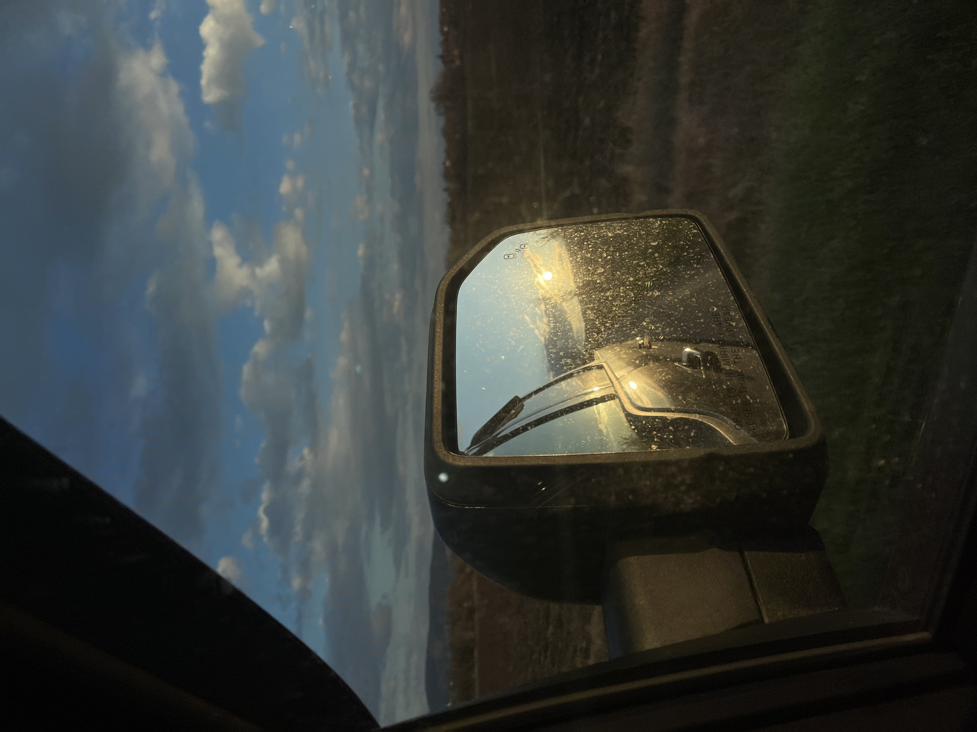 The image shows a car side mirror reflecting a landscape. The sky is visible with clouds and a hint of sunlight. Part of another car is reflected in the mirror. The environment is outside and somewhat blurry. There is no indication of the location being Yardzhilovtsi, Bulgaria.