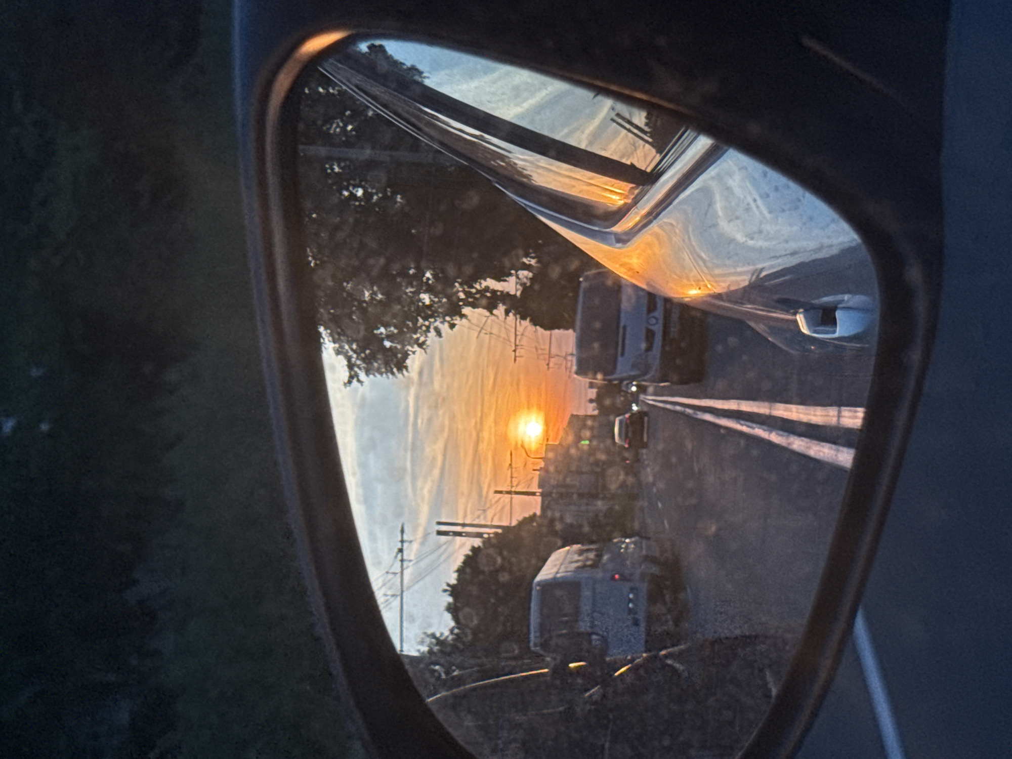 The image is a reflection in a car's side-view mirror, capturing a sunset over a rural road. The setting appears to be Vinnytsia, Ukraine, based on the provided location context. The sun is low on the horizon, casting a warm, orange glow across the sky, which is streaked with wispy clouds. The road is flanked by trees and utility poles, suggesting a semi-rural or suburban environment.
Several vehicles are visible on the road, including what appear to be campers or RVs, suggesting a journey or a leisurely drive. The reflections in the mirror are somewhat distorted, as is typical of this type of photograph. The overall mood is peaceful and serene, with the setting sun creating a beautiful and atmospheric scene. There is no visible text in the image. The time of day is clearly sunset, and the weather appears clear with some cloud cover. The location cues like the trees and utility poles, combined with the vehicles and the general landscape, point towards a journey on a road outside of a major urban center.
