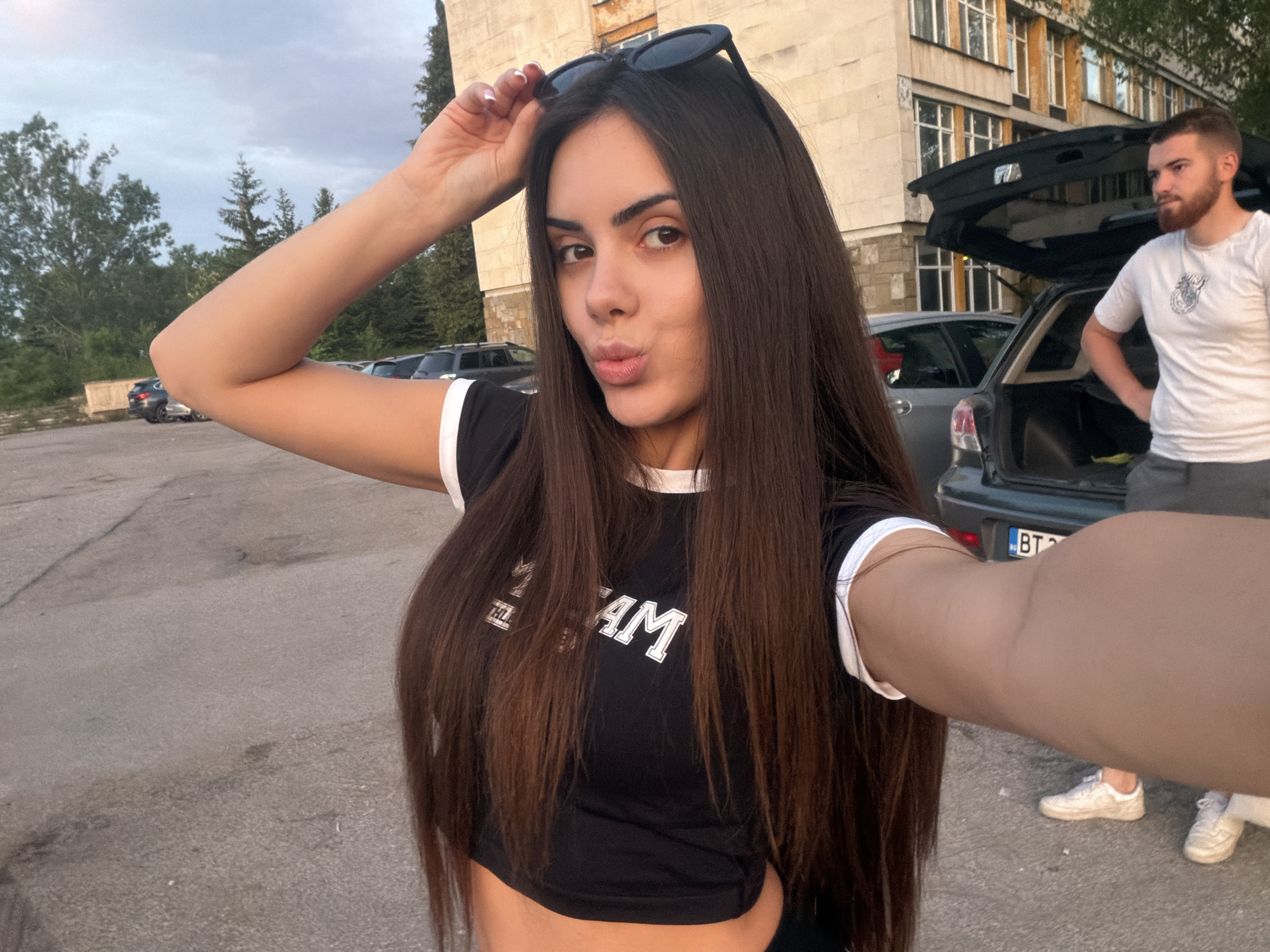 A young woman takes a selfie outdoors. She has long, straight, brown hair and is wearing a black crop top with white trim. She is holding sunglasses on top of her head and puckering her lips for the photo. A man stands in the background, near a car with its trunk open. The car has a Bulgarian license plate, reading "BT." The setting appears to be a parking lot near a building with multiple windows. Trees can be seen in the distance. Based on the license plate and building style, the location may be the city of Sofia, Bulgaria.