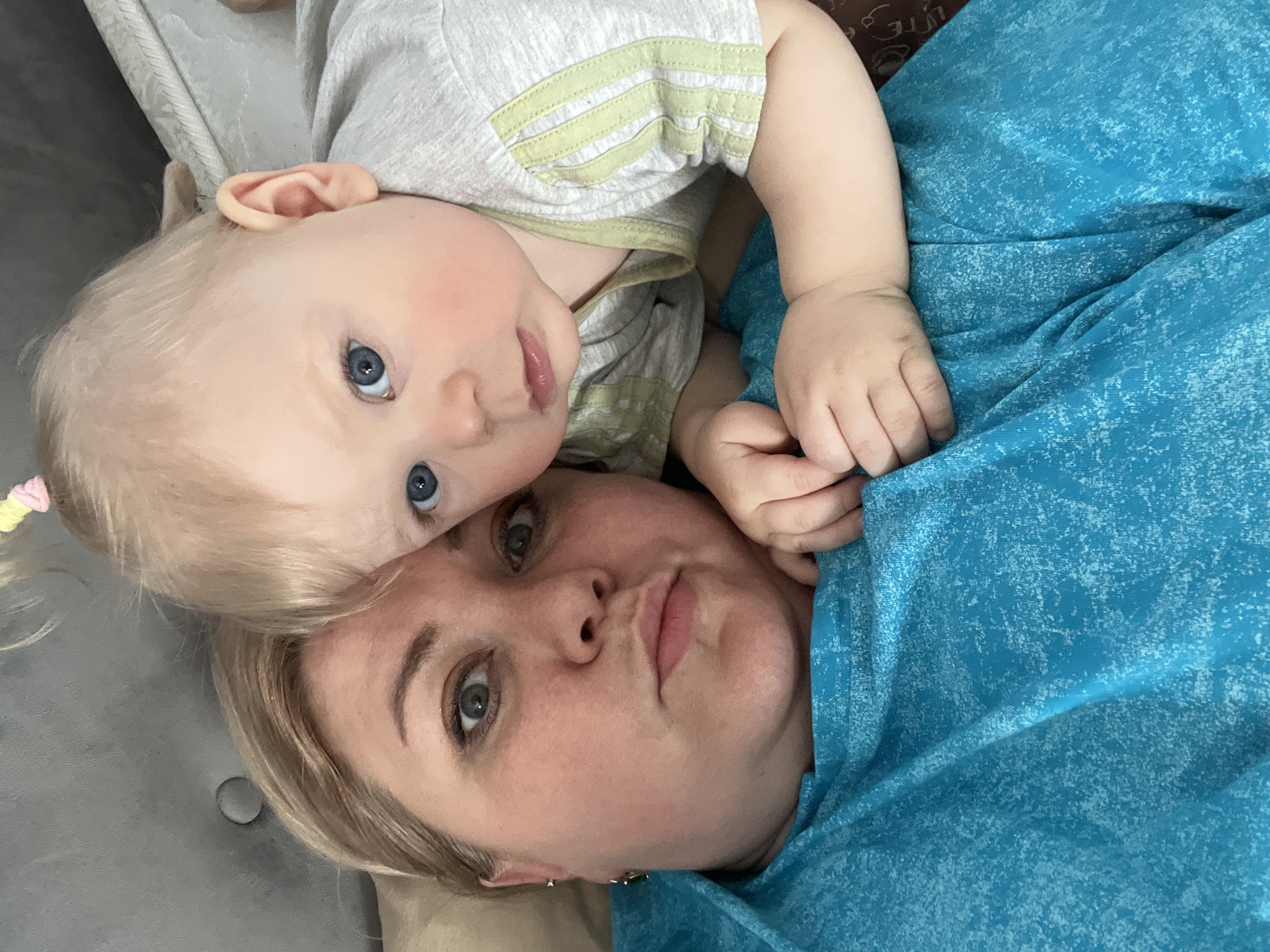 This image, taken in Vesyoloye, Russia, features a woman and a baby, likely mother and child, lying down together, potentially on a sofa or bed, capturing a close-up selfie. The image is oriented sideways, with both subjects looking directly at the camera.
The woman, positioned at the bottom, has blonde hair, light-colored eyes (blue-grey), and fair skin. She is wearing a vibrant teal or blue top with a subtle, lighter-colored speckled pattern. Her lips are slightly pursed in a playful pout. Visible in her ear is a dark green earring.
The baby, positioned above the woman's head, has fair skin, blonde hair, and striking blue eyes. The baby is wearing a light grey short-sleeved top with lime green horizontal stripes on what appears to be a chest pocket. One of the baby's hands is visible near the woman's shoulder.
The background consists of a light grey, textured surface, possibly upholstery. The lighting suggests it is daytime. There is no visible text in the image.