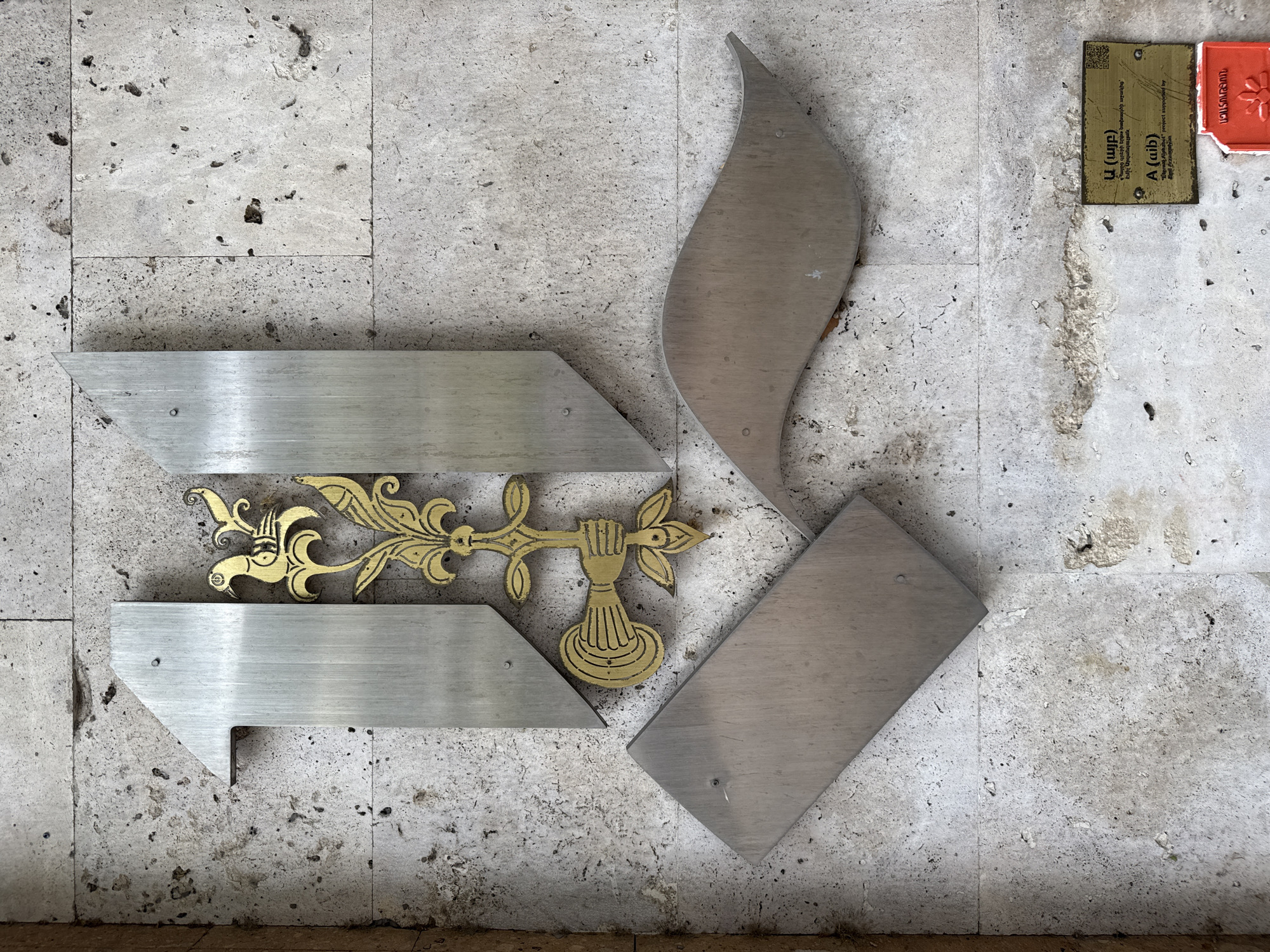 The image shows a segment of a stone wall with metallic artistic elements. A large, stylized "1" is prominently displayed on the left, made of brushed metal. To its right, a decorative gold element features a bird perched on a branch with floral motifs. Below the "1", a rectangular metallic plate is attached to the wall. To the right of the gold decoration, a sweeping, curved metallic element is present. At the bottom right of the image, a small informational plaque with text in Armenian and English is affixed to the wall. The plaque explains that the artwork is part of the "Mesrop Mashtots Alphabet Project" and is supported by the Arzoumanian family. The surrounding wall is constructed of light-colored, textured stone tiles with visible grout lines and some natural imperfections. The scene appears to be an outdoor setting in the city of Yerevan, Armenia.
