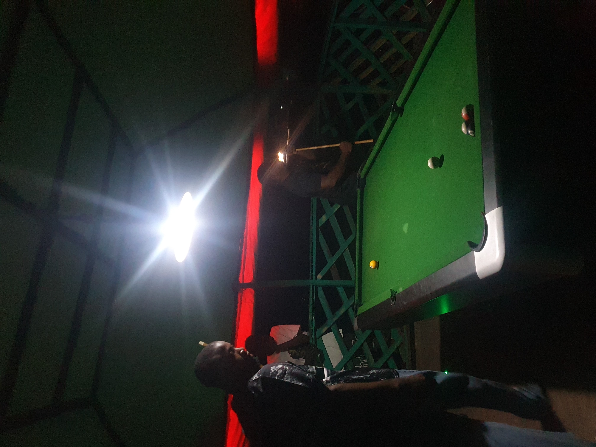 In Lokoja, Nigeria, two men are playing pool under a green, tent-like roof with red fabric draped behind them. A bright light fixture illuminates the scene, creating lens flare. One man, wearing a black shirt, is in the foreground, looking down at the pool table. Another man, in a dark shirt, stands further back, holding a cue stick and appearing to be about to strike a ball. The pool table is green, with several balls on it, including a yellow ball, a white ball, and a cluster of three balls, one of which appears to be red and white striped. A green lattice fence is visible behind the pool table. The overall atmosphere is dimly lit, suggesting an outdoor or semi-outdoor recreational space.