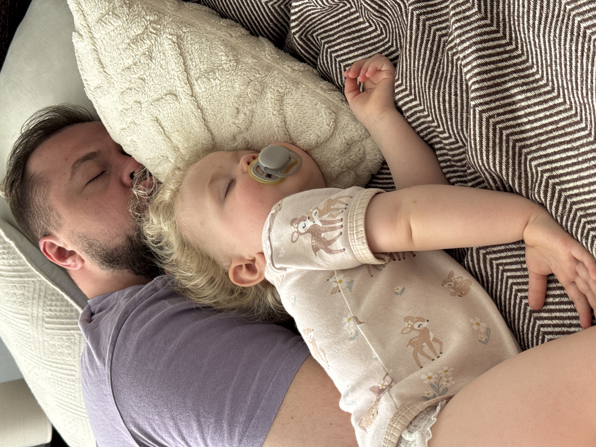 The image shows a man and a baby sleeping on a bed. The man, with a beard and a grey t-shirt, is lying on his back with his eyes closed. The baby, wearing a patterned onesie and with a pacifier in its mouth, is lying on top of him. A textured pillow is beneath their heads, and a brown and white chevron-patterned blanket covers part of the scene. The image was not taken in Vladimir, Russia.