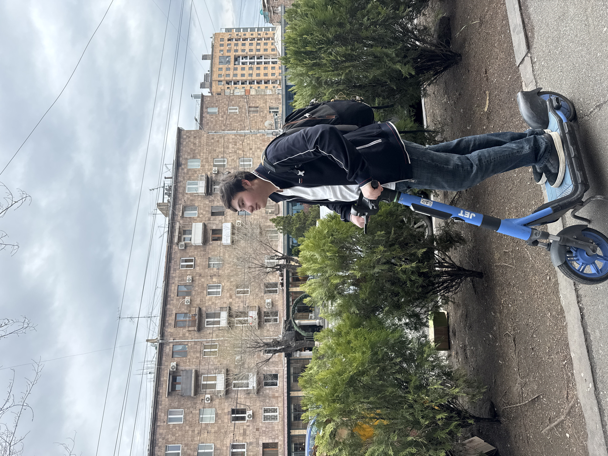 A young man rides a blue "JET" branded scooter on a sidewalk in an urban setting. He wears a black jacket with white stripes, jeans, and sneakers, and carries a black backpack. The background features a row of apartment buildings with visible air conditioning units and power lines. Evergreen bushes are planted along the sidewalk. The sky is overcast. The city is labeled as Unknown, Unknown.