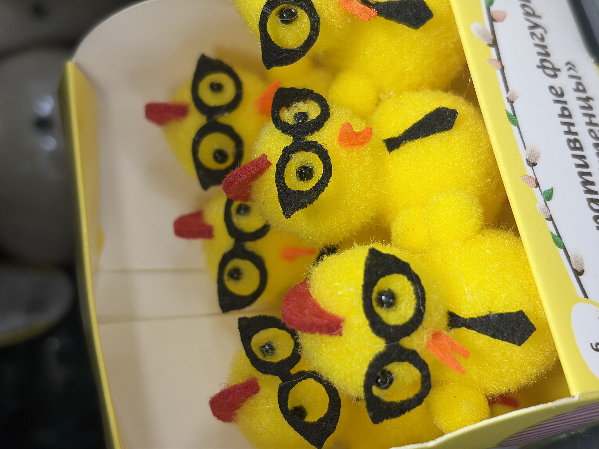 A box of yellow toy chicks sits inside a store, likely in Saint Petersburg, Russia, based on the Russian text printed on the box. The chicks are fluffy, bright yellow, and each has a set of black-rimmed eyes, along with a small orange or red beak. A black tail is also visible on some. The box appears to be made of cardboard and is predominantly yellow with a white interior. The writing on the box is partially visible. There is no visible human presence in the image. The environment outside the box is blurry, but it suggests a store setting with other merchandise nearby.