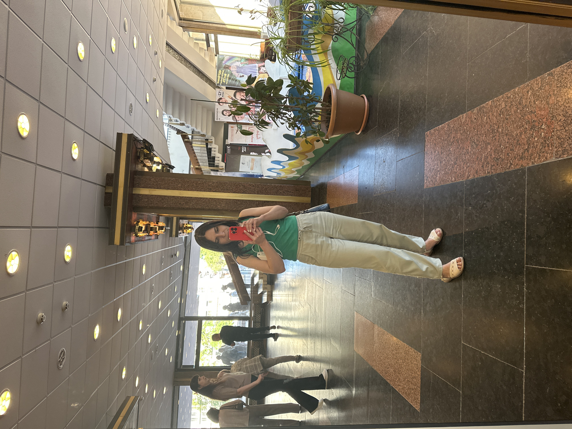 The image displays an indoor public space, likely a lobby or concourse, in the city of Unknown, Unknown. The primary subject is a woman, reflected in a large mirror or reflective wall on the right side of the frame, taking a selfie. She has dark hair and wears a green tank top, light beige drawstring pants, and light-colored flat sandals with woven straps. She holds a red smartphone with a visible multi-lens camera in both hands, oriented towards herself, with white wired earbuds dangling from it. A small white object is clutched in her left hand, and a blue strap is visible over her left shoulder.
In the reflection behind her, other individuals are visible: a woman in a light-colored short-sleeve top and dark pants walking left, another person in a light plaid shirt, and a man in a dark shirt walking away from the viewer. Large windows along the left side and background of the reflected space reveal an outdoor urban environment with trees and buildings.
The interior features a high ceiling composed of white acoustic tiles with numerous circular recessed lights, some emitting a warm yellow glow and others clear white light. Several small white smoke detectors or vents are also present on the ceiling. Ornate columns with gold or bronze decorative trim and polished reddish-brown stone cladding are prominent architectural elements. On the right side, a light-colored staircase with dark handrails and balusters ascends.
In the mid-ground right, a low, wavy decorative counter or wall is painted in bright segments of blue, green, yellow, and white. In front of this counter, a potted plant with dark green leaves sits in a terracotta pot on the dark-tiled floor. To its right, another potted plant rests on an ornate black metal stand. Behind these elements, multiple large posters featuring human faces and text in an unknown script are visible.
The floor throughout the area is comprised of large dark gray or black rectangular tiles, interspersed with narrower reddish-brown rectangular strips of granite or similar stone, forming pathways.