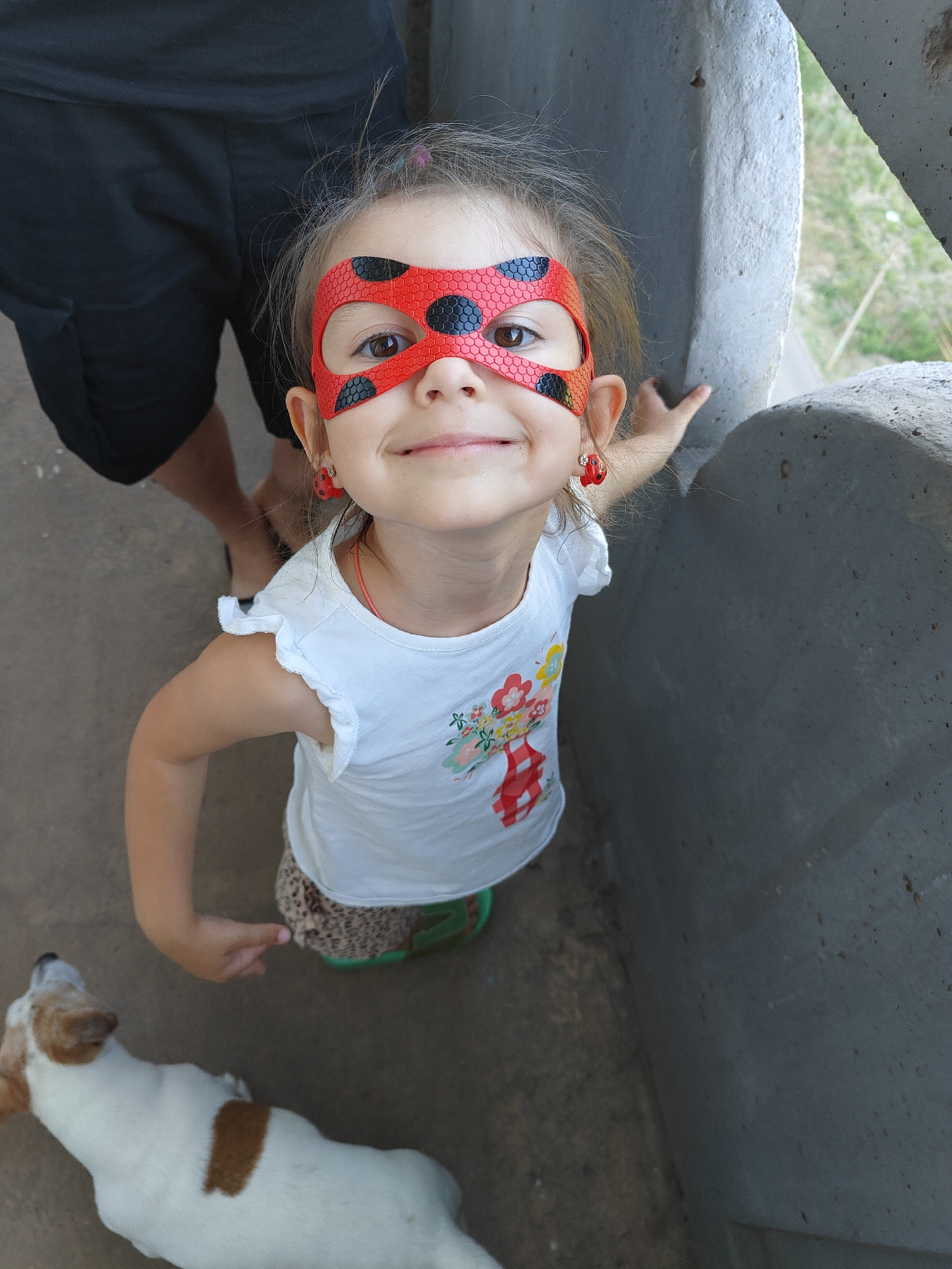 A young girl wearing a red and black ladybug mask and matching earrings smiles at the camera. She has a white t-shirt with floral and checkered patterns, and leopard-print shorts with green shoes. She is standing outdoors, with a concrete structure behind her and a dog in the foreground. A person in black shorts stands to the left. The scene is captured from a high angle.