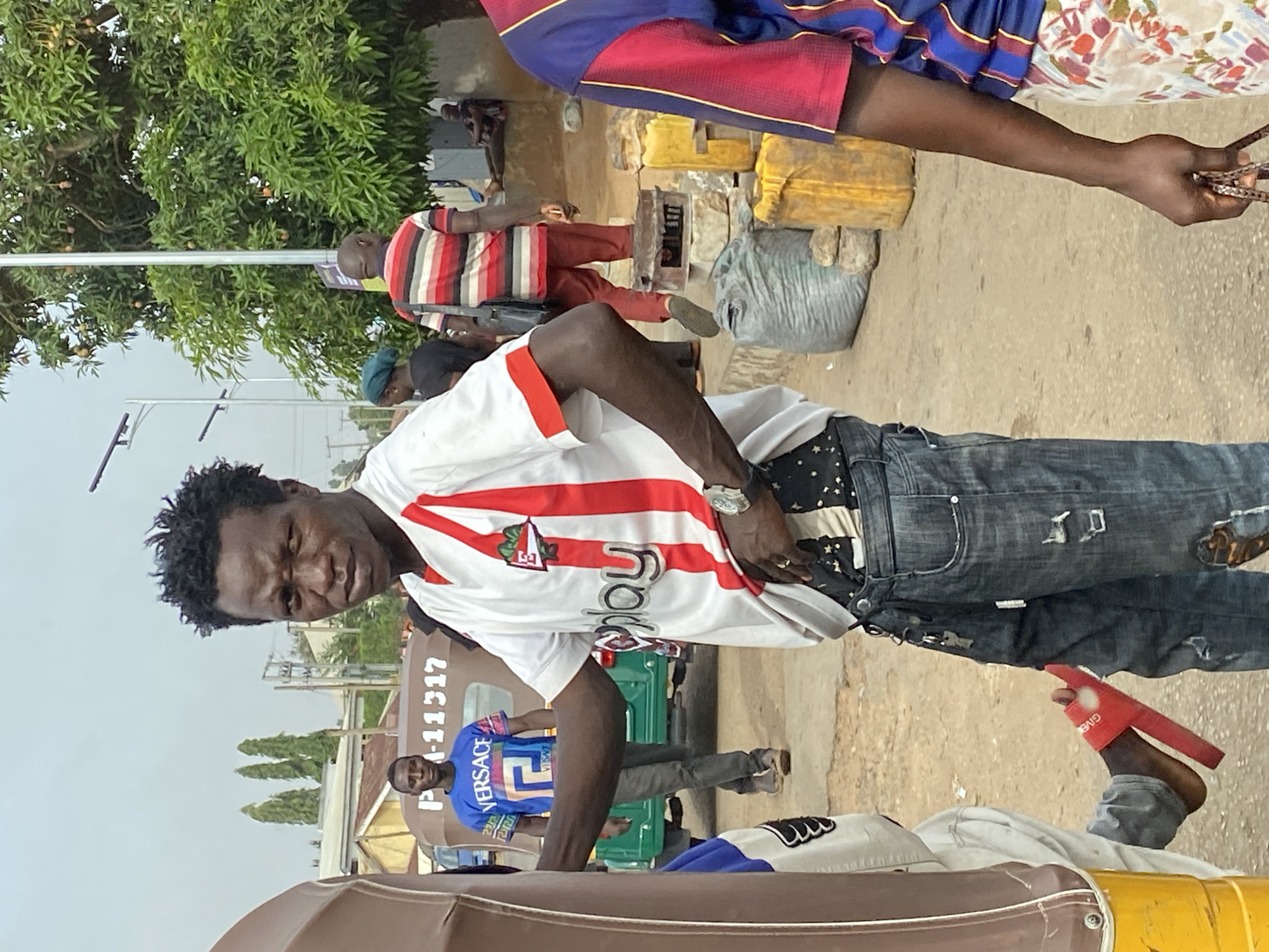 The image captures a street scene in Bukuru, Nigeria. A man wearing a white shirt with red stripes and distressed jeans is prominently featured, with a wristwatch visible on his left wrist. Other people are visible in the background, including a man in a blue Versace shirt standing near a green vehicle displaying the license plate P-11317. In the distance, people appear to be selling items on the side of the road, with yellow objects (possibly food items) and a grey sack present. The setting is outdoors, with trees lining the background and a light-colored sky overhead. A section of a brown vehicle is visible in the lower foreground.