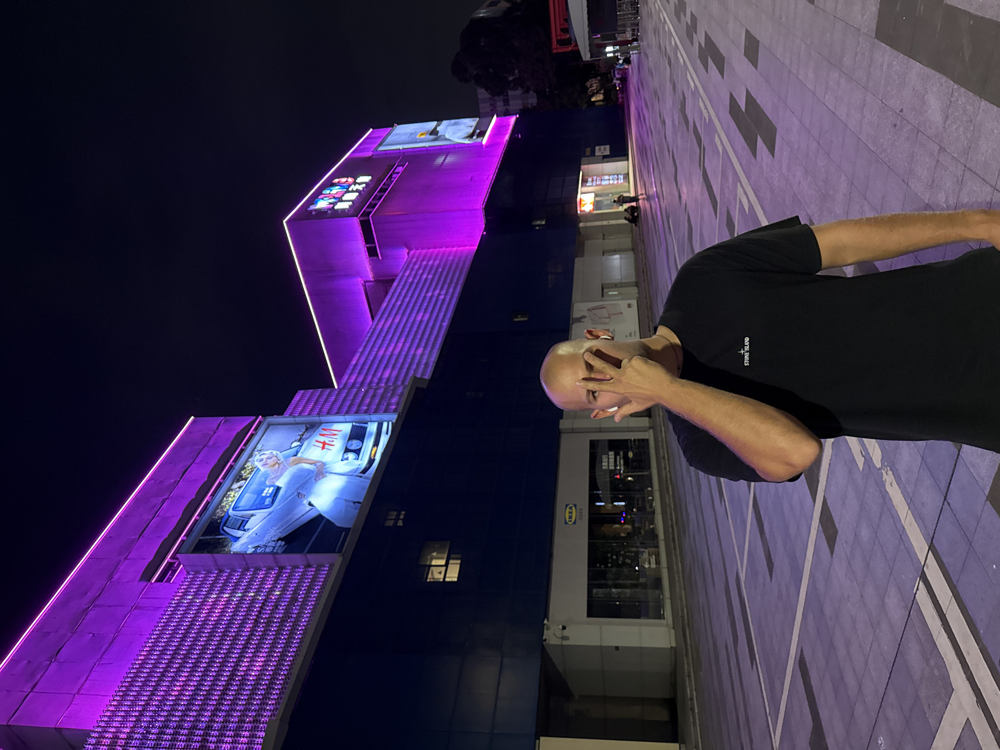 The image depicts a vibrant night scene in a modern urban environment, likely a commercial district. The setting is dominated by large, multi-story buildings illuminated with striking purple and pink neon and LED lights against a dark night sky. The ground is paved with large, light-colored square and rectangular tiles, forming a pattern with darker sections and white lines.
In the foreground, on the right side of the frame, a man is partially visible. He is bald and wearing a black t-shirt that features a small white "STONE ISLAND" logo on the left chest. His left hand is raised to his face, making a gesture that covers part of his face, while his right arm is visible by his side. He appears to be posing for the camera.
The architectural style of the buildings is contemporary. The main building on the left is extensively lit with purple/pink lights outlining its structure and features. Several large digital billboards are integrated into its facade. One prominent screen on the lower left displays an advertisement for "H&M", showing a woman in white sitting on the hood of a white car. Another screen higher up shows a grid of colorful icons or displays, and a third, further back, shows a light-colored advertisement.
Behind the man, another building features large glass windows, and a distinctive "IKEA" logo in blue and yellow is clearly visible, accompanied by Chinese characters. This detail, along with the overall modern city appearance, confirms the location in China, specifically Tianhe District as per context. The scene is brightly lit by artificial lights, indicating nighttime.