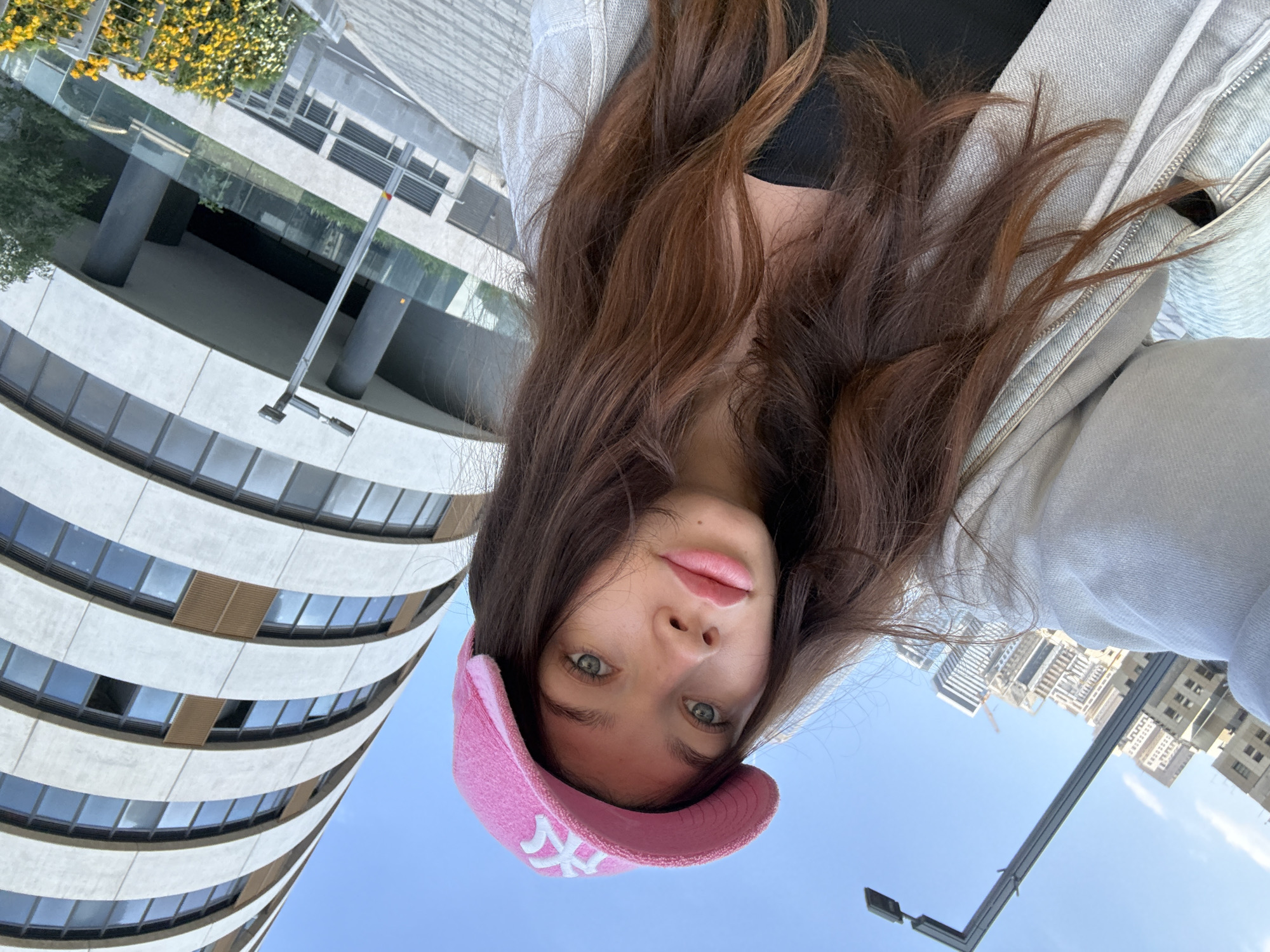 A young woman with long, wavy brown hair and prominent green eyes is captured in a close-up, low-angle shot, looking directly into the camera. She has full, naturally pink lips and a small beauty mark above her upper lip. She is wearing a bright pink baseball cap with a white "NY" logo on the front, along with a light-colored, possibly light grey, hooded jacket over a dark top.
The setting is an outdoor urban environment, likely Tel-Aviv, Israel, under a clear, bright blue sky, indicating daytime. Behind and above the woman, a modern architectural structure dominates the left side of the frame. This building features a curved facade with multiple horizontal bands of windows and light-colored panels. Near the top left, some lush yellow and white flowering plants add a touch of greenery. On the right side of the background, other contemporary high-rise buildings are visible, some appearing to be under construction, with a faint crane outline. A grey street light pole extends into the frame from the right side, adding to the urban landscape. The overall atmosphere suggests a sunny and clear day in a bustling city. The woman's expression is neutral and direct, suggesting a posed moment.