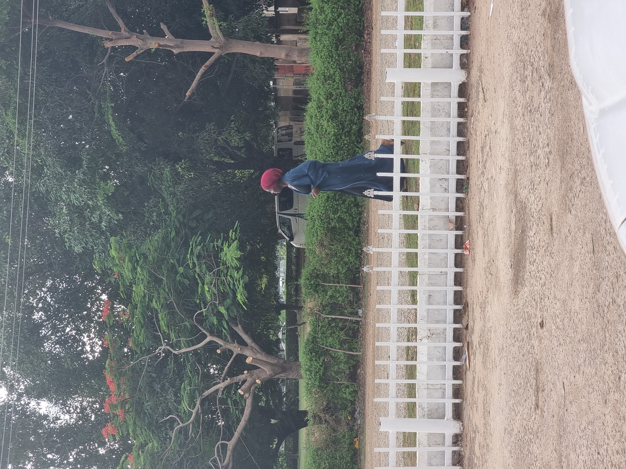 An elderly individual wearing a red cap and a blue robe is walking near a white metal fence in Gwagwalada, Nigeria. The scene includes trees and a green hedge behind the fence. A vehicle is partially visible in the background. The ground is a sandy color.