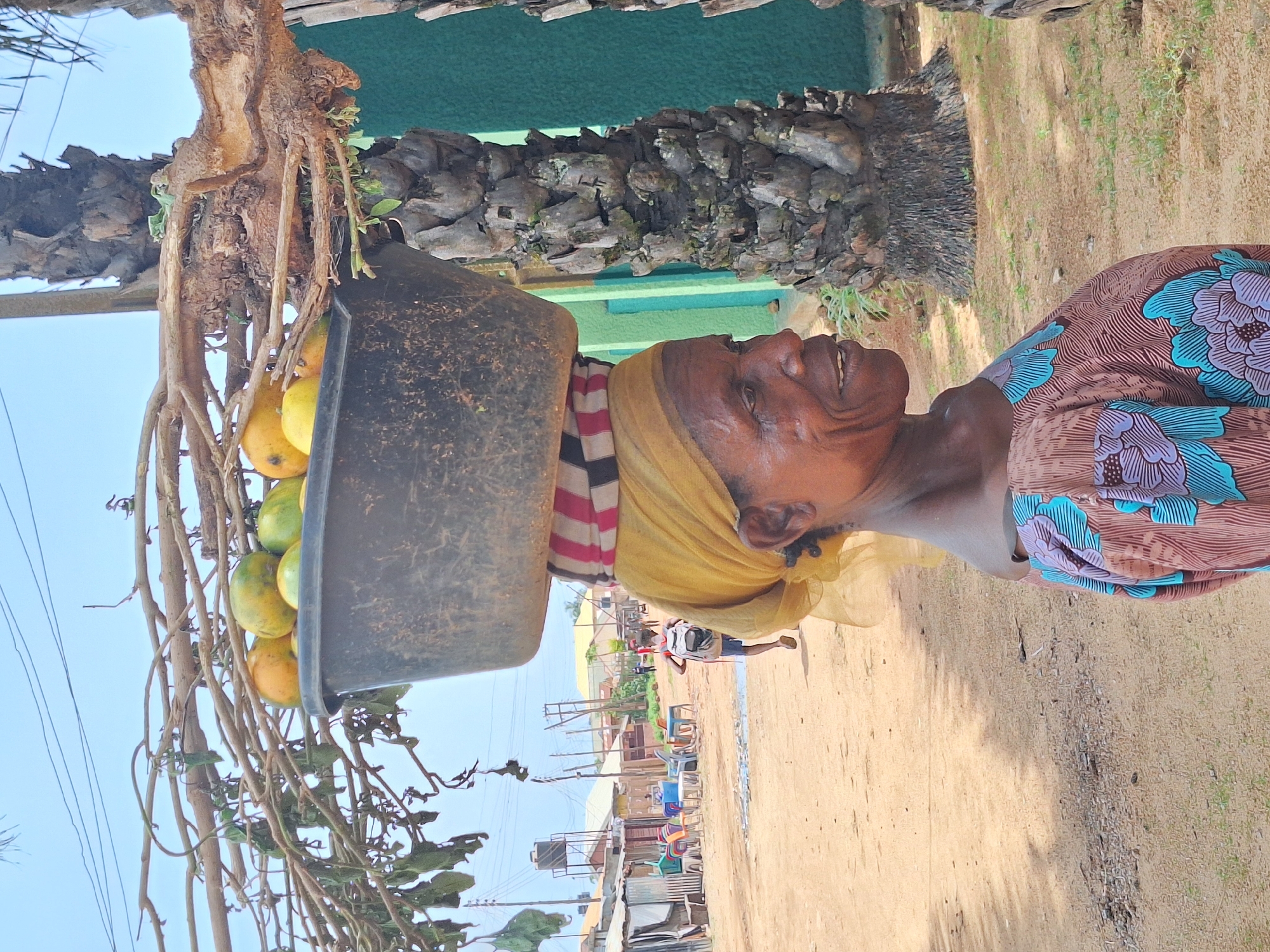 An elderly woman in Sabon Tasha, Nigeria, carries a black rectangular tub filled with mangoes atop her head. She wears a yellow headscarf, a striped cushion beneath the tub, and a brown and blue floral print top. The tub is positioned on a network of branches. She is walking along a sandy road. Behind her, there are buildings, utility poles, and other people in the distance. A section of a palm tree is also visible near the woman.