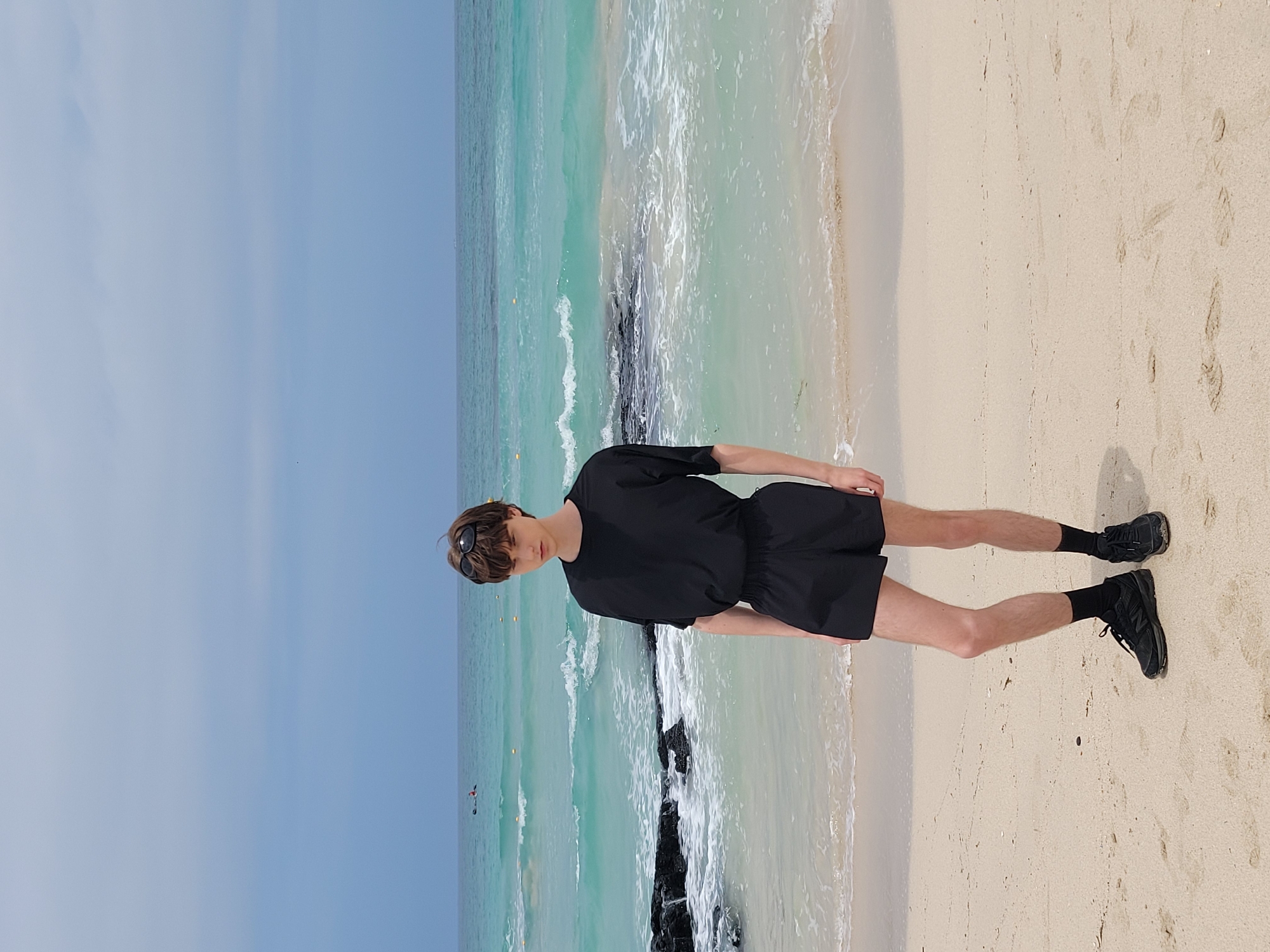 A young person stands on a sandy beach with the ocean in the background. They are wearing a black t-shirt, black shorts, black socks, and black sneakers. Sunglasses are perched on their head. The ocean is a bright turquoise color with white waves crashing on the shore. There are dark rocks visible in the water on the left side of the image. The sky is overcast with light gray clouds. The scene appears to be a natural, coastal environment. This location is likely a beach in Jeju, South Korea, known for its beautiful turquoise waters and volcanic rock formations.