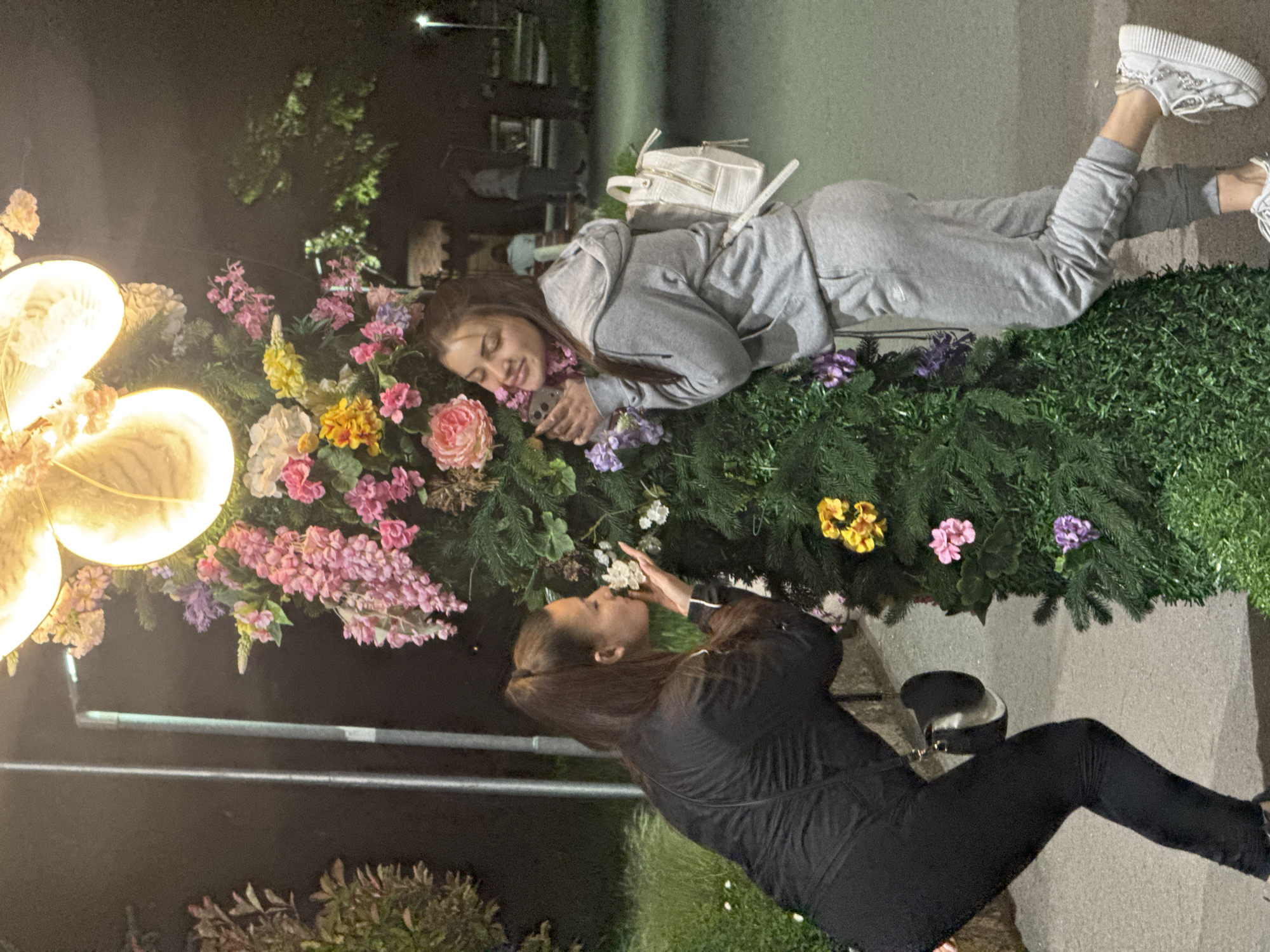 Two young women are interacting next to a flower bed illuminated by a large artificial flower light feature in Shumen, Bulgaria. One woman, dressed in a grey tracksuit and white sneakers, is lying on the flower bed, seemingly looking at her phone. The other woman, dressed in black athletic wear, is kneeling on the ground, seemingly either smelling or admiring the flowers. The environment appears to be a public space at night, with benches and other people faintly visible in the background. There are trees and grass visible. The flowers in the bed include pink, yellow, and white varieties, surrounded by greenery. A white backpack is resting on the grey-clad woman.
