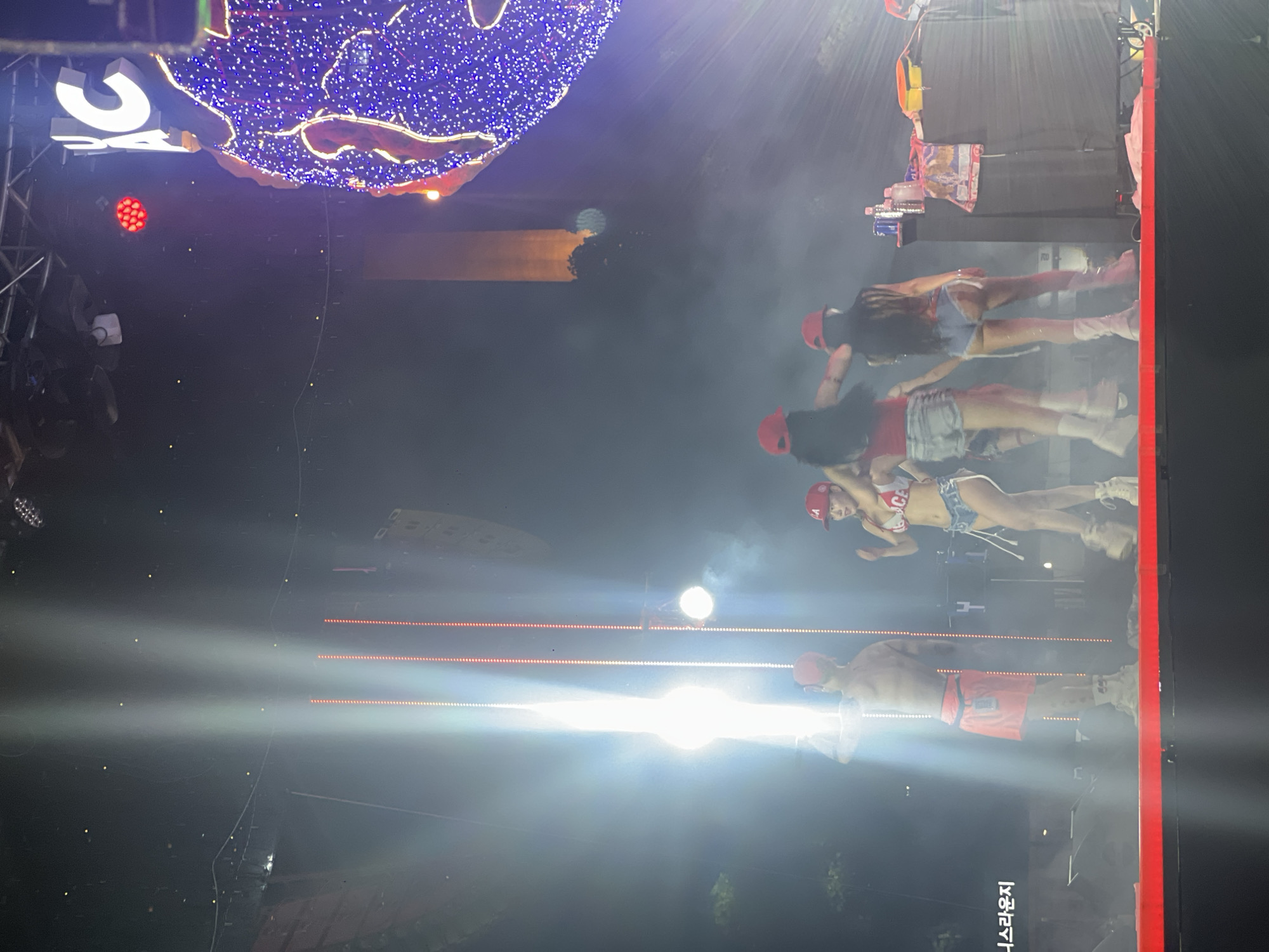 The image captures a night-time outdoor event, possibly a concert or festival, in Daegu, South Korea. A brightly lit stage is the central focus, with intense white spotlights casting beams into the smoky atmosphere. On the stage, several individuals are performing. A shirtless male performer, wearing a red skirt and white sneakers, stands prominently under a strong spotlight, his back to the camera. To his right, three female performers in red tops and denim shorts are also on stage, one in the foreground facing the camera and two with their backs to the camera. The performers appear to be engaging in a dance or performance. In the background, large speakers and lighting equipment are visible, along with a partially obscured, large illuminated structure, possibly a decorative art installation or part of the stage design, adorned with blue and yellow lights. The overall scene suggests a lively and energetic event.