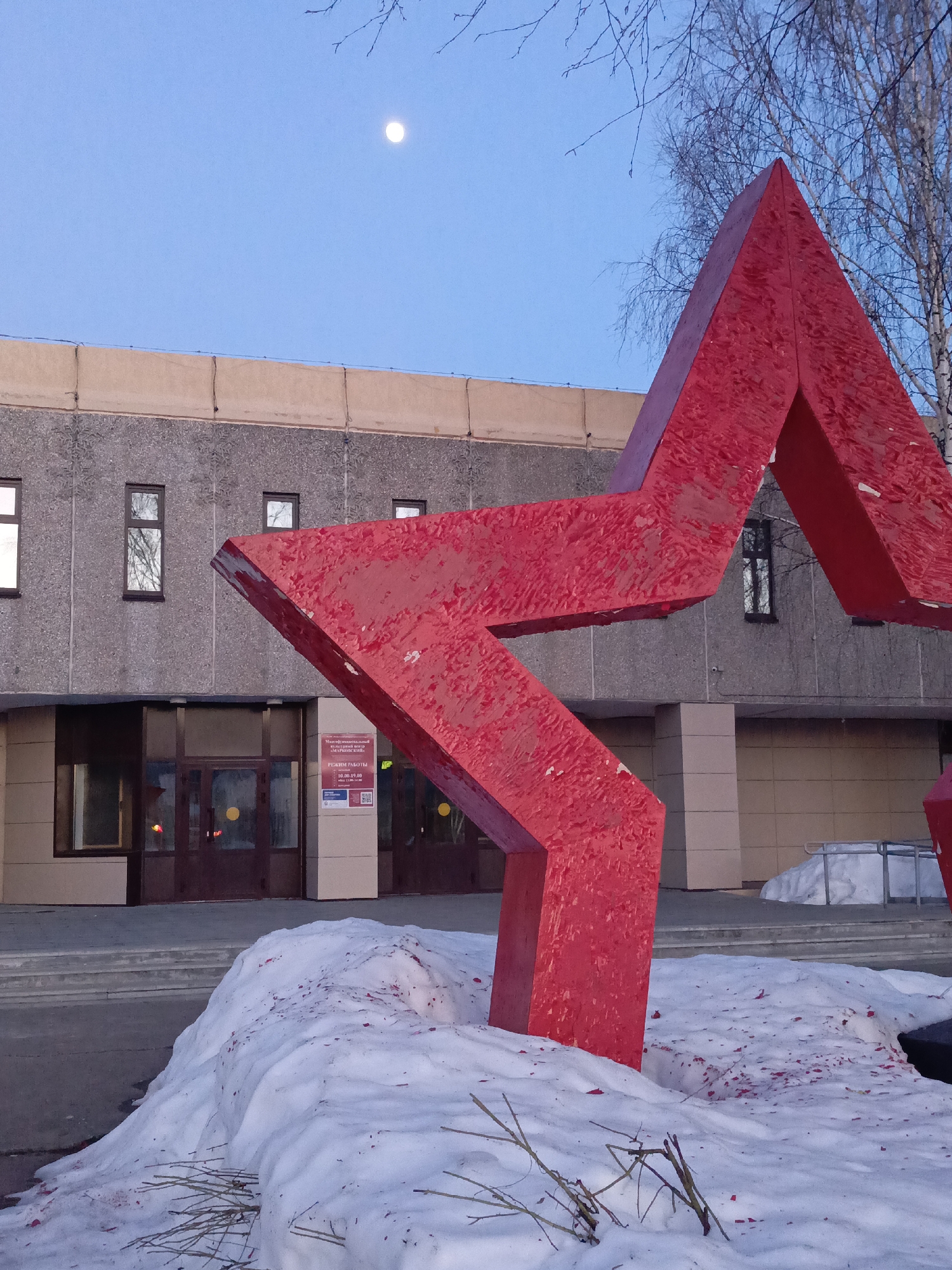 The photograph depicts a large, red, five-pointed star statue partially covered in snow, located in front of a building in Марковский, Russia. The building behind the statue is two stories tall and has a concrete facade with rectangular windows and a glass-paneled entryway. A sign with text in Russian is visible above the entrance. The ground is covered in a mix of snow and bare pavement. A bare tree is visible to the right of the statue. A full moon is visible in the light blue sky above the building. The statue appears to be coated in red paint with some chipping and wear. The snow around the base of the statue is also speckled with red paint.