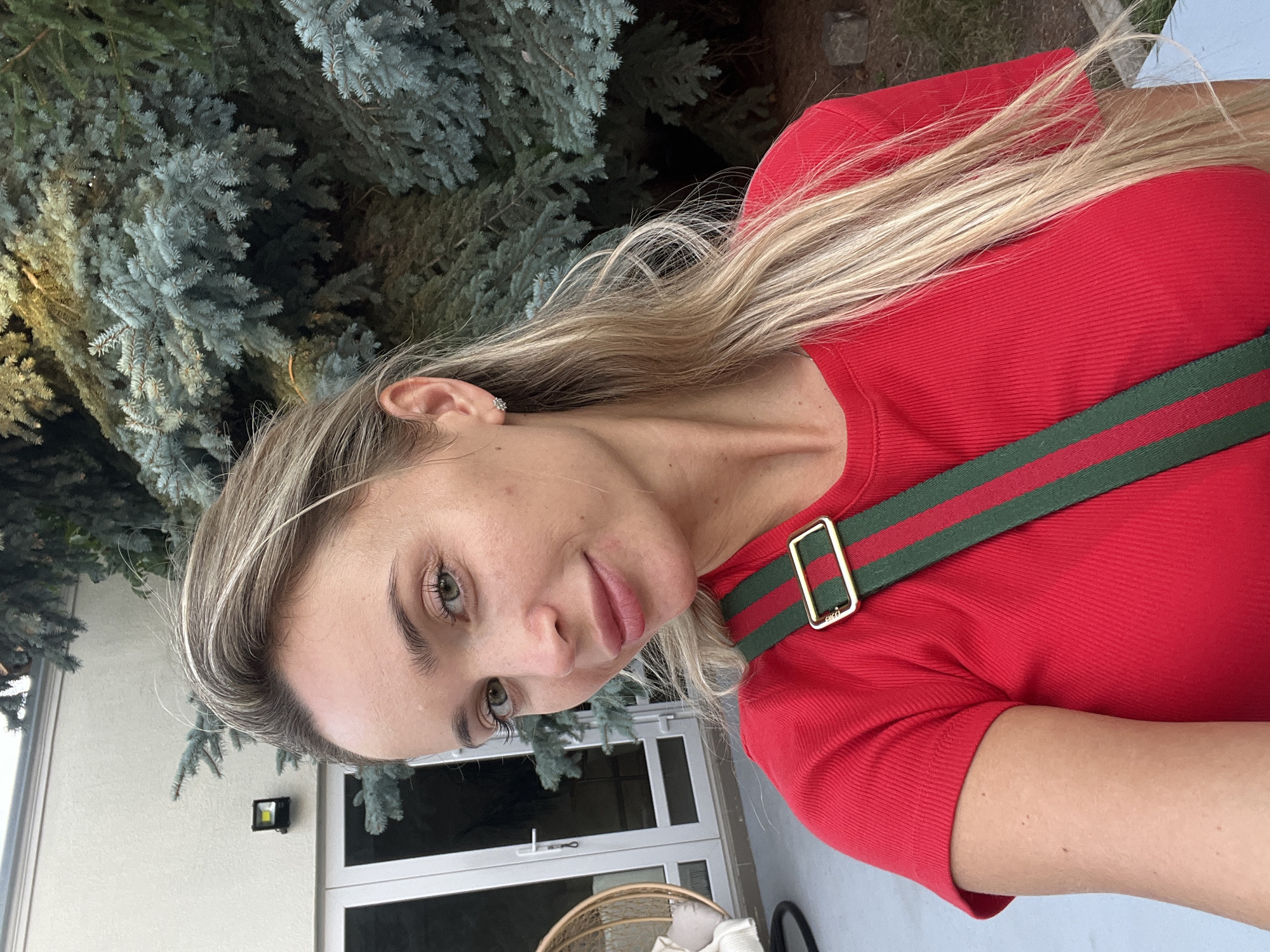 A woman with blonde hair and green eyes is taking a selfie outdoors. She is wearing a red ribbed t-shirt with a green and red striped strap across her chest. In the background, there is a light-colored building with large glass doors, a hanging chair, and lush green trees. The scene appears to be in a residential area or a garden. There is no indication of the location being in the city of Fontanka, Ukraine.