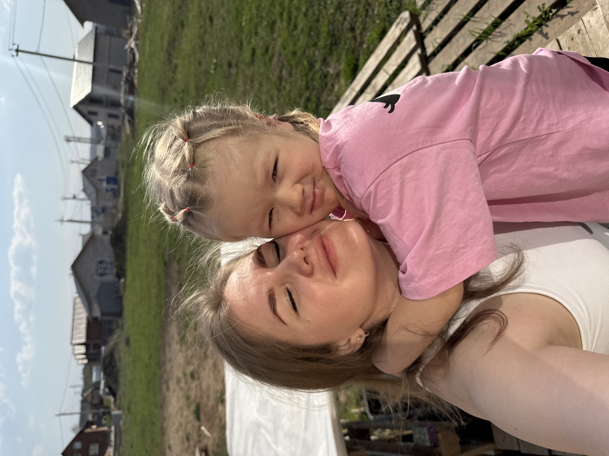 A mother and child are outdoors in Ферма, Russia. The mother, with brown hair and wearing a white tank top, lies on her back. The child, wearing a pink shirt, lies on top of her, embracing her. In the background are houses, green grass, and a blue sky. A wooden bench is visible.